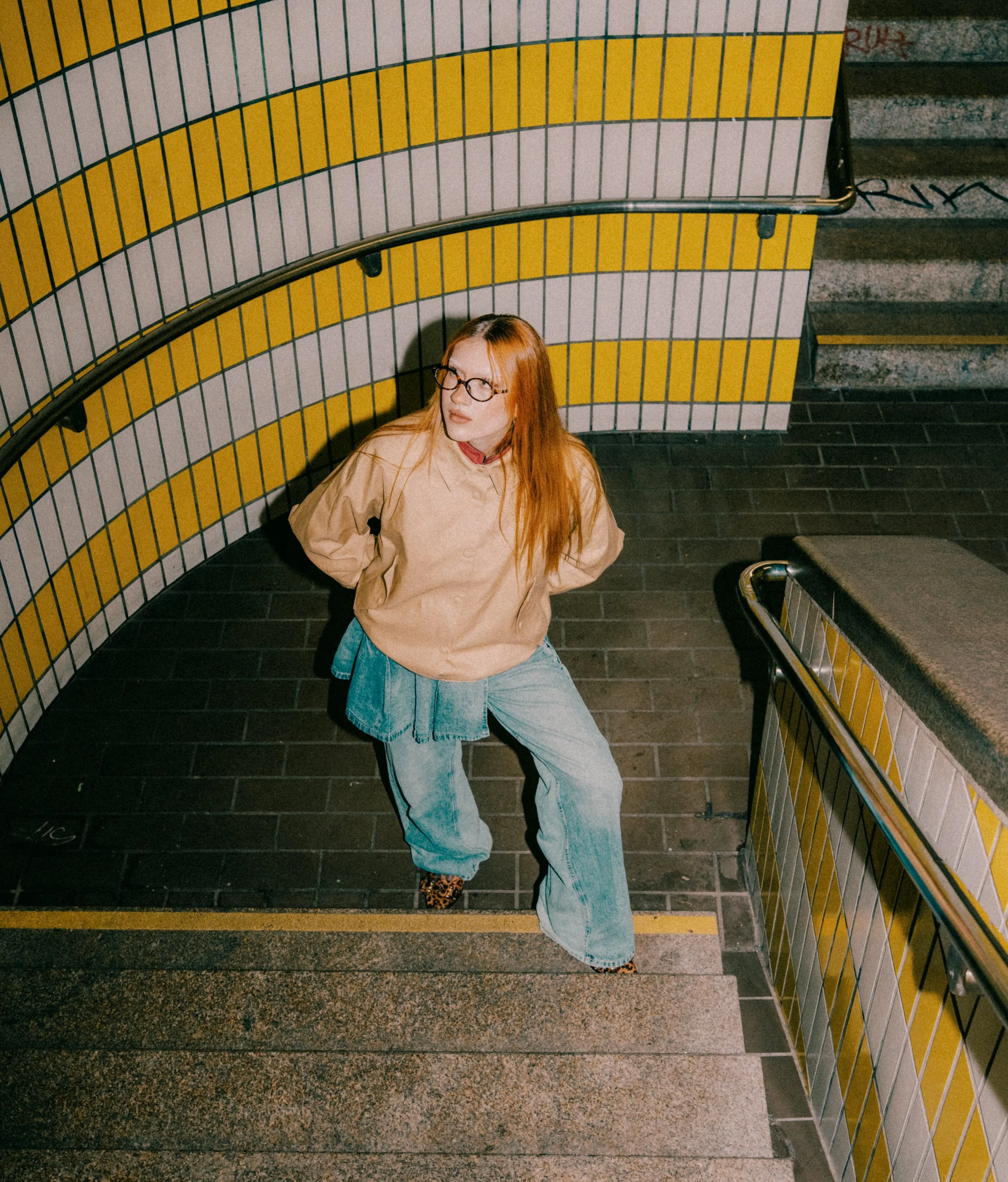 Eine Frau mit roten Haaren, runden Brillen und bunten Schuhen sitzt an einer Treppe in einer U-Bahn-Station, umgeben von gelb-weißer Kachelwand.