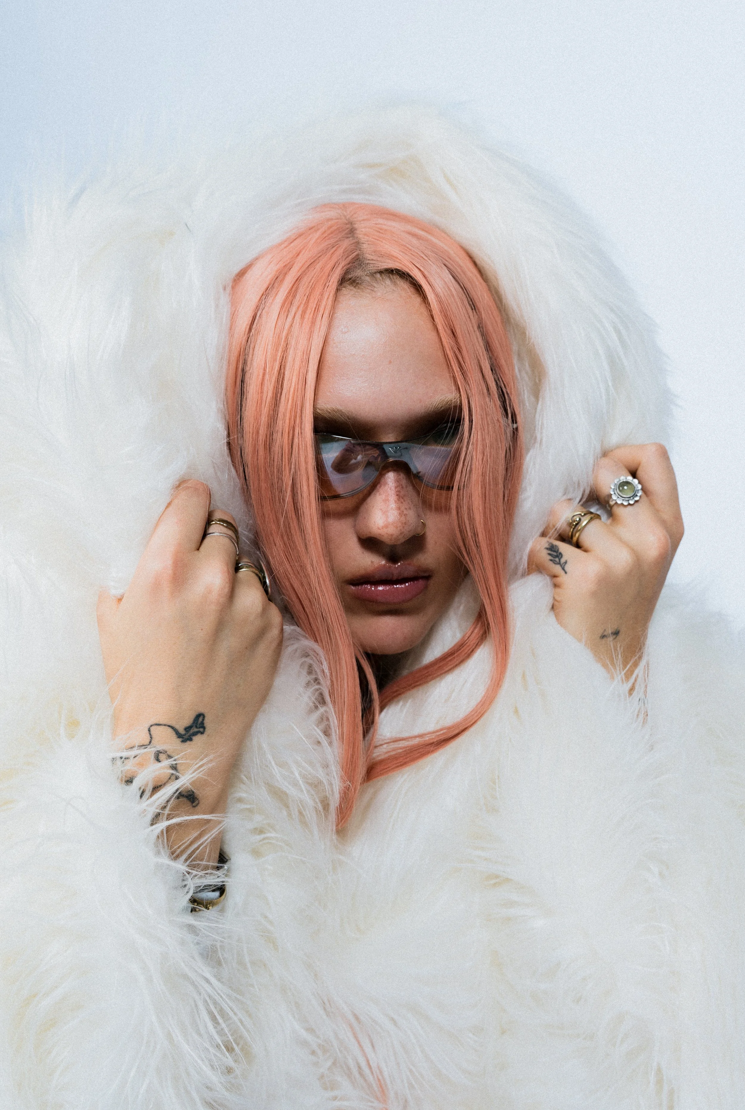 Junge Frau mit rosa Haaren, dunklen Sonnenbrillen, in einer weißen Felljacke, mit Tattoo auf Hand, mehrere Ringe, posiert vor einem weißen Hintergrund.