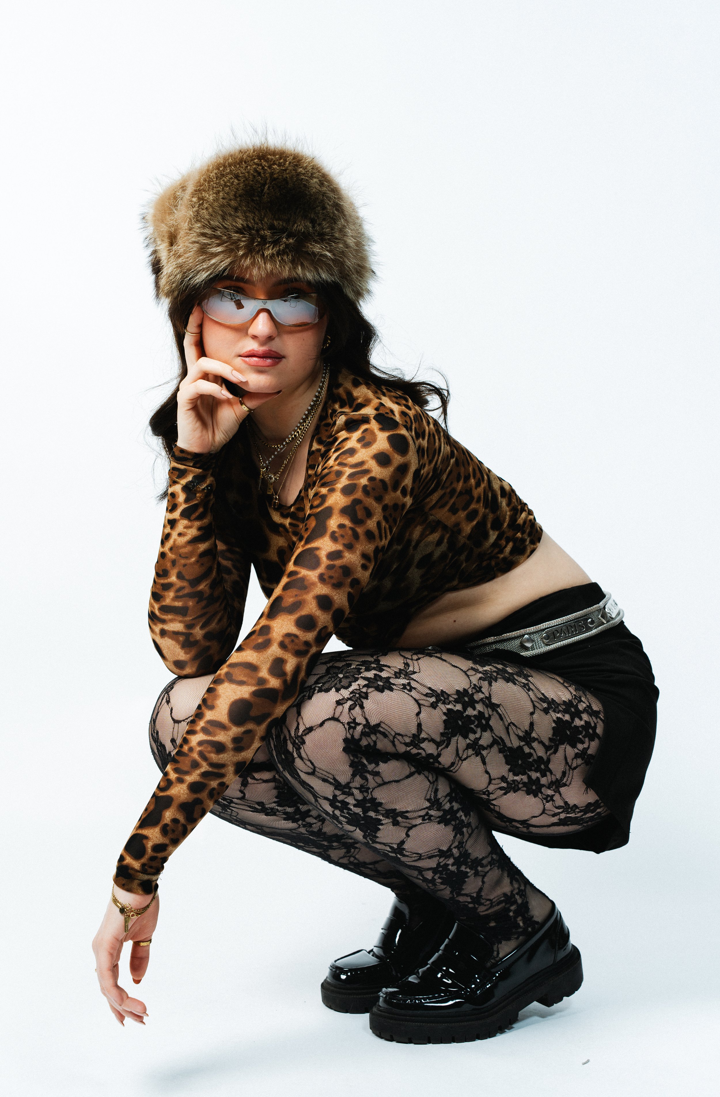 Junge Frau in Animal-Print Kleidung, mit Fellmütze, Sonnenbrille, Strumpfhosen mit Spitzenmuster, schwarze Schuhe, posiert gegen weißen Hintergrund.