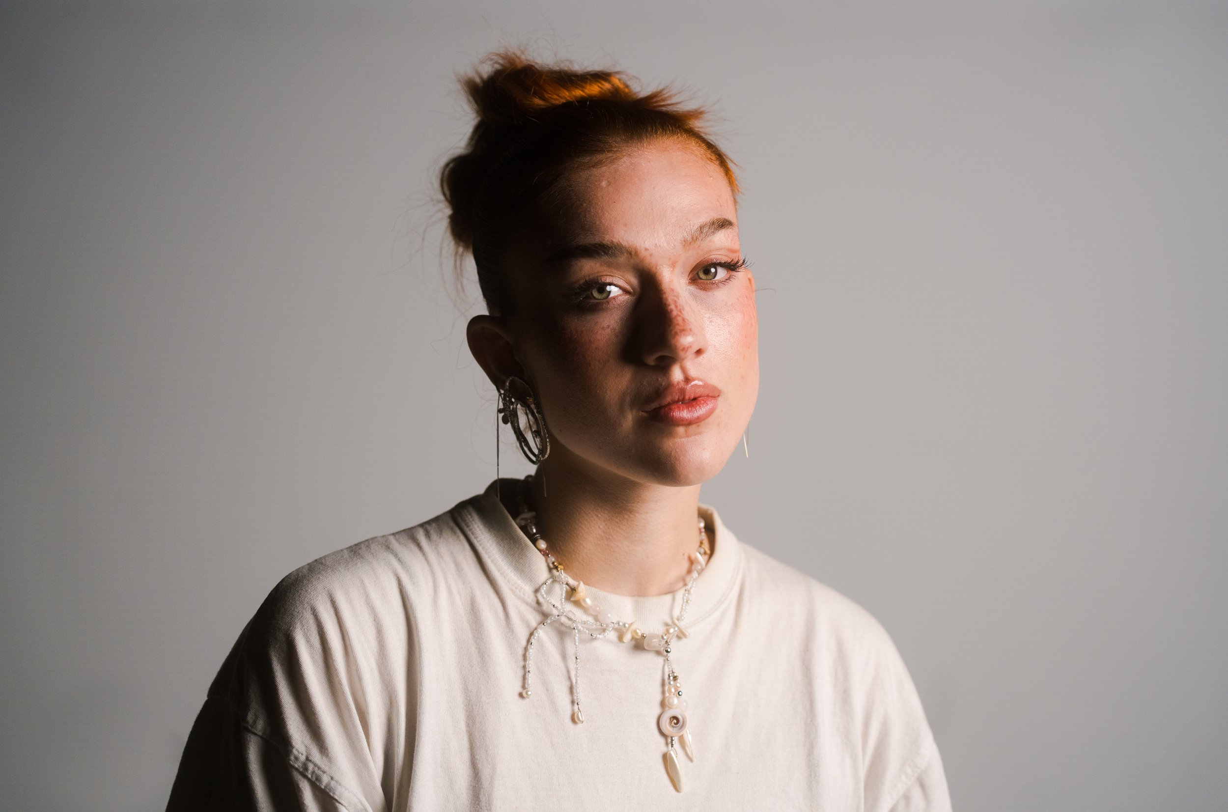 Porträt einer jungen Frau mit roten Haaren, die einen neutralen Ausdruck zeigt, getragen mit mehreren Schmuckstücken, vor einem grauen Hintergrund.