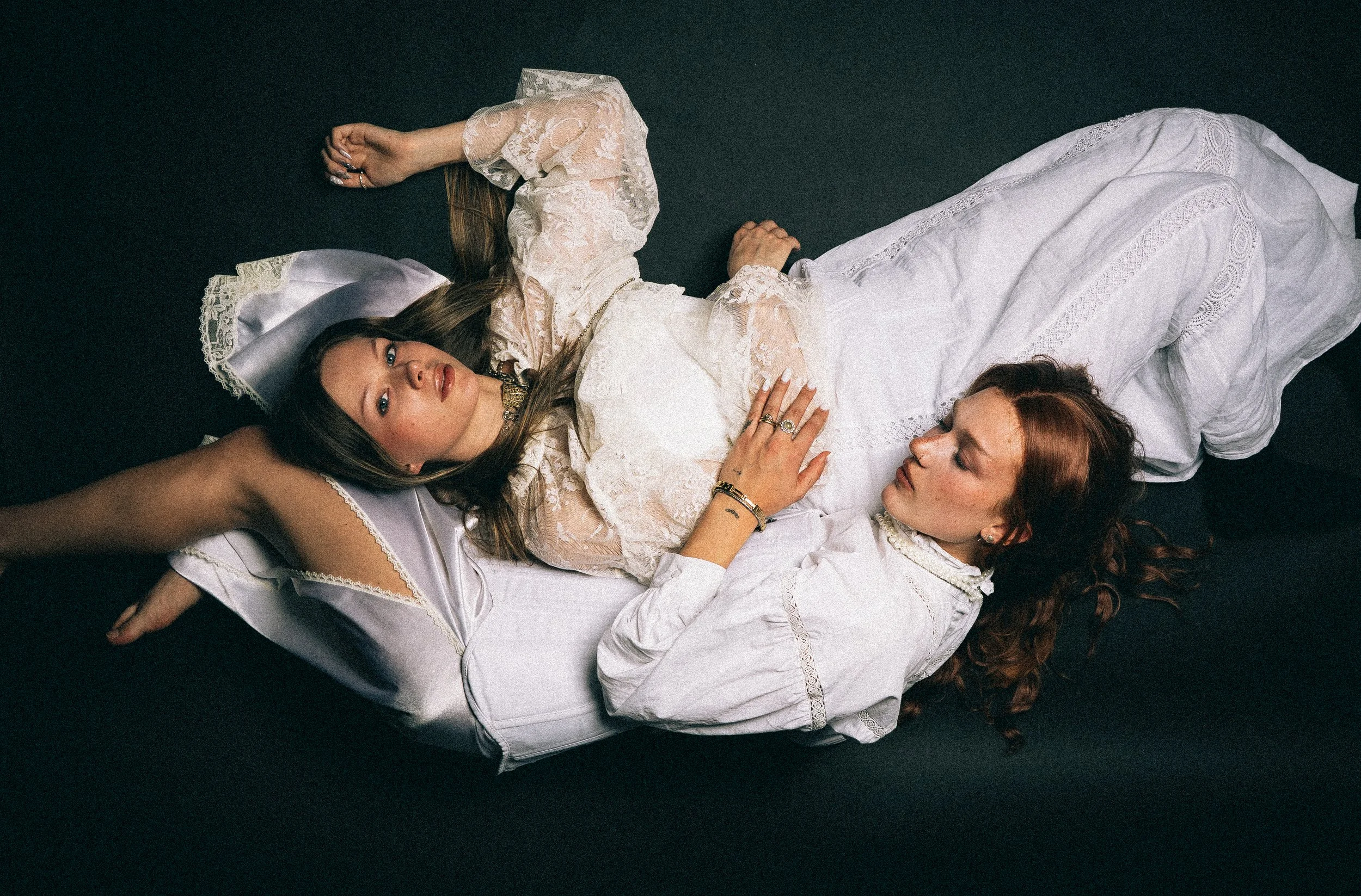 Zwei Frauen liegen auf dem Boden, die eine auf dem Rücken, die andere seitlich, und schauen in die Kamera. Beide tragen weiße, romantische Kleidung mit Spitzen. Das Bild hat einen dunklen Hintergrund.