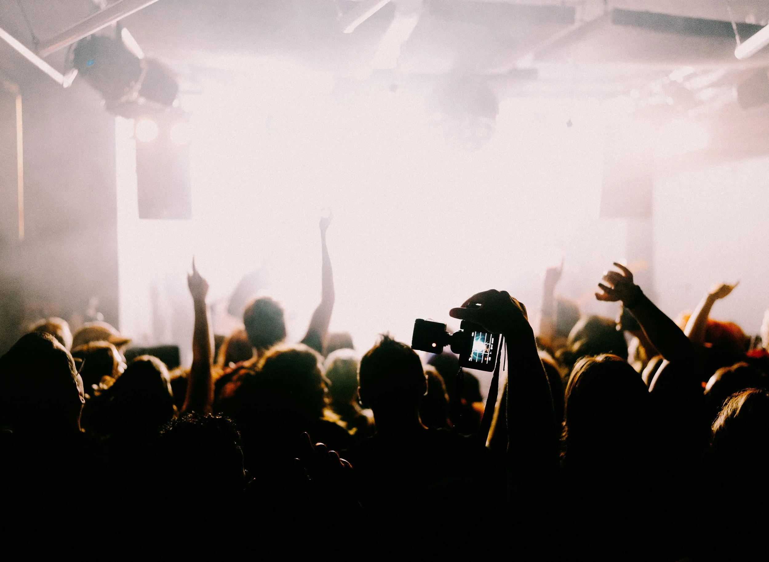 Menschenmenge bei einem Konzert oder einer Veranstaltung, einige Personen fotografieren oder filmen die Bühne, Licht- und Tontechnik sichtbar, dunkle Silhouetten vor hellem Hintergrund.