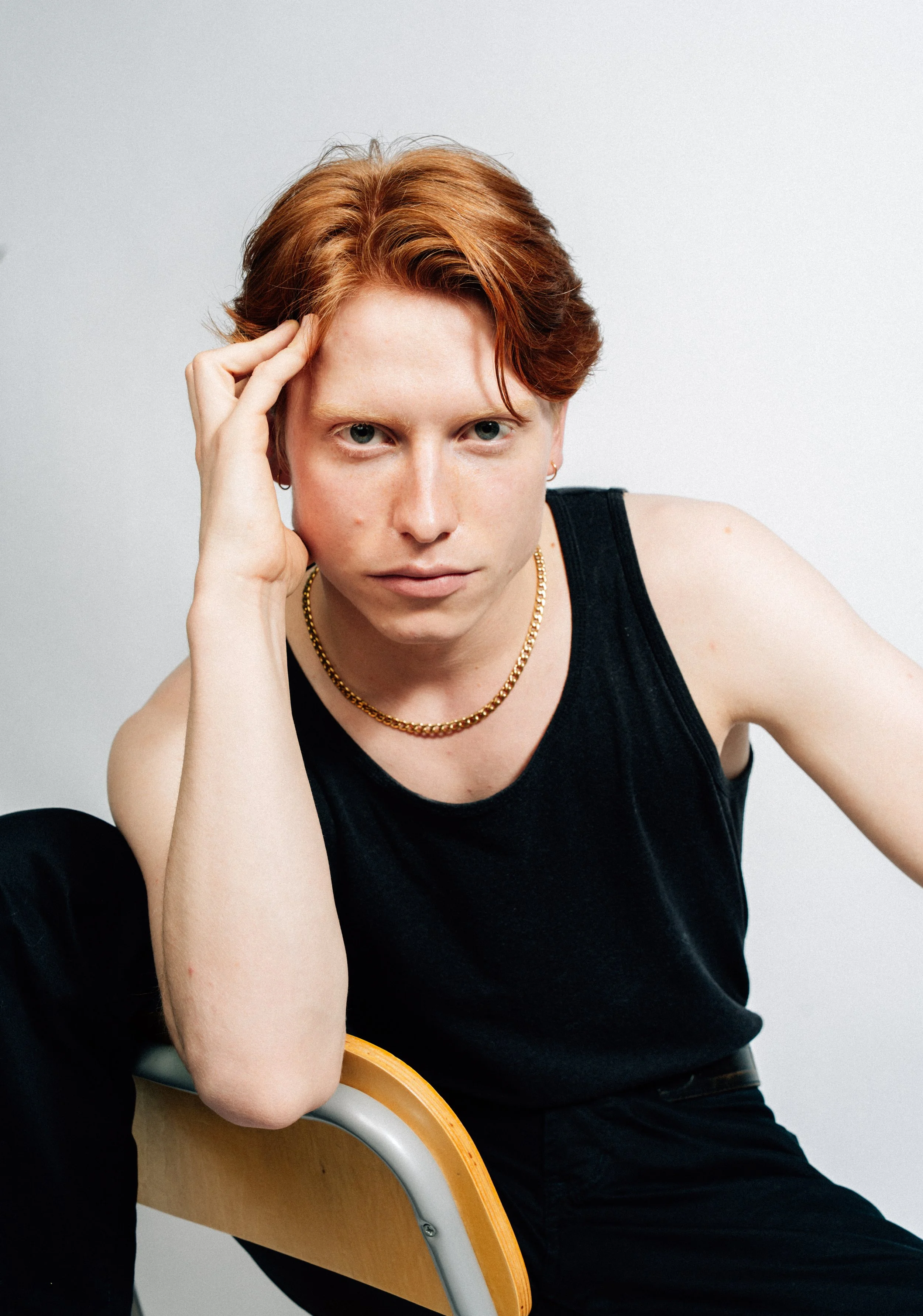Porträt eines jungen Mannes mit roten Haaren, schwarzen Kleidung und Goldschmuck, sitzend auf einem Stuhl gegen eine weiße Wand.
