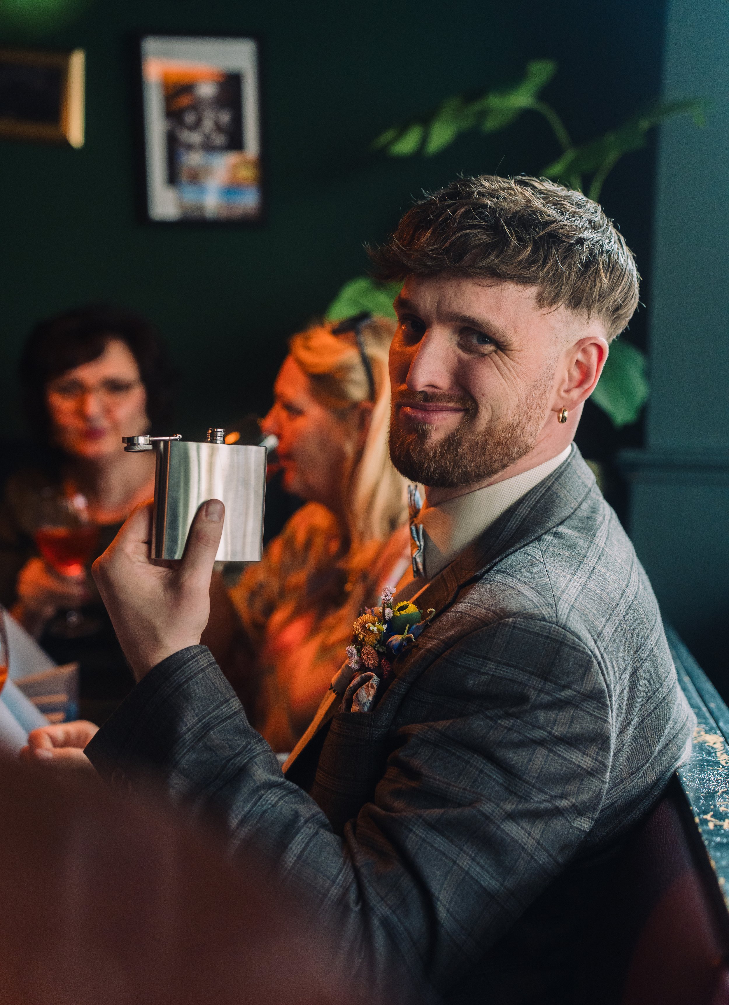 Ein junger Mann mit grauem Anzug und Blumen im Revers hält ein Flachmann und lächelt in die Kamera, während im Hintergrund drei Frauen in einer geselligen Runde sitzen, Wein trinken und sprechen.