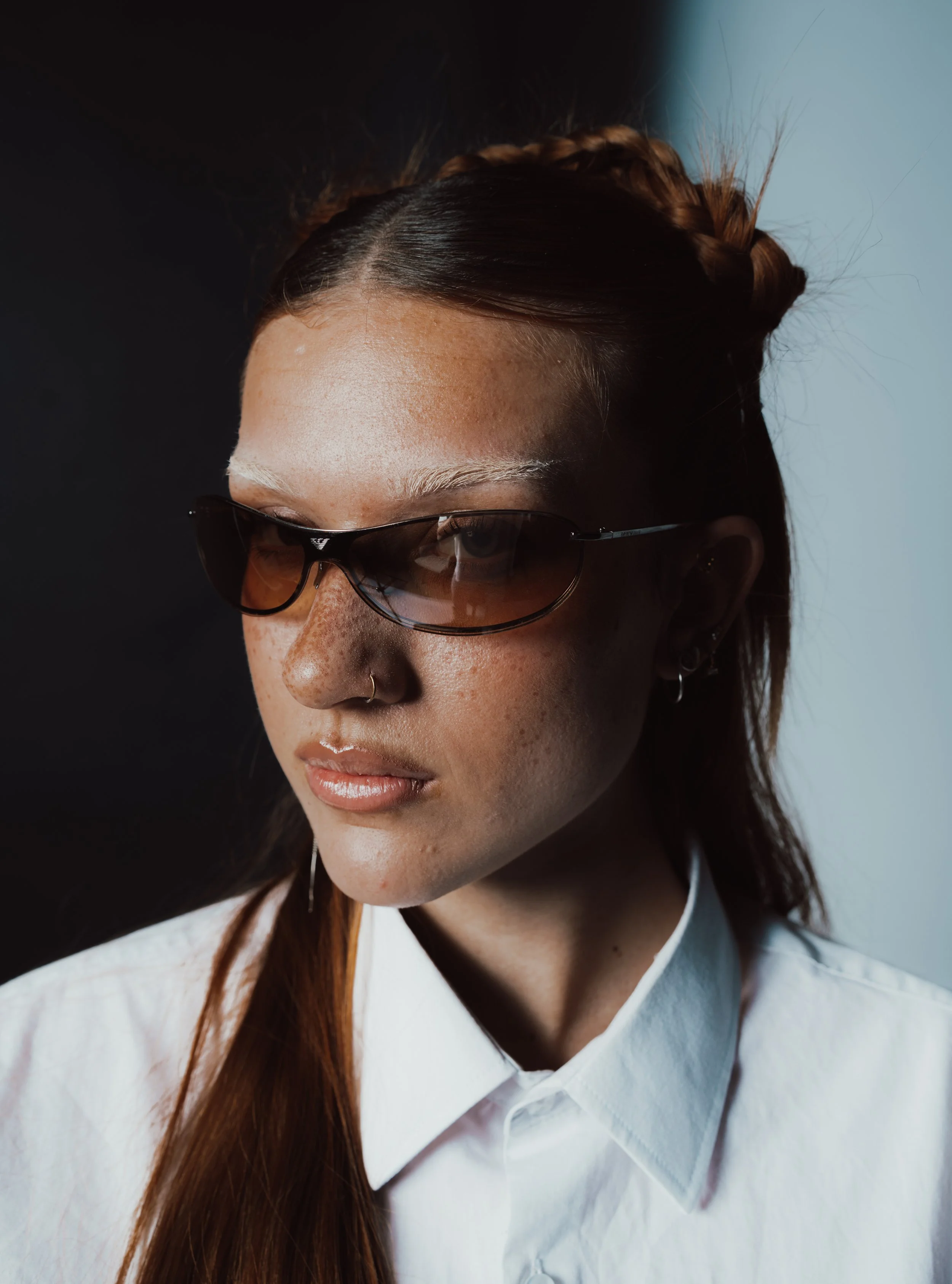 Porträt einer jungen Frau mit Sonnenbrille, hellblonder Haut, Locken und weißen Hemd vor einem geteilten Hintergrund aus Schwarz und Hellblau.