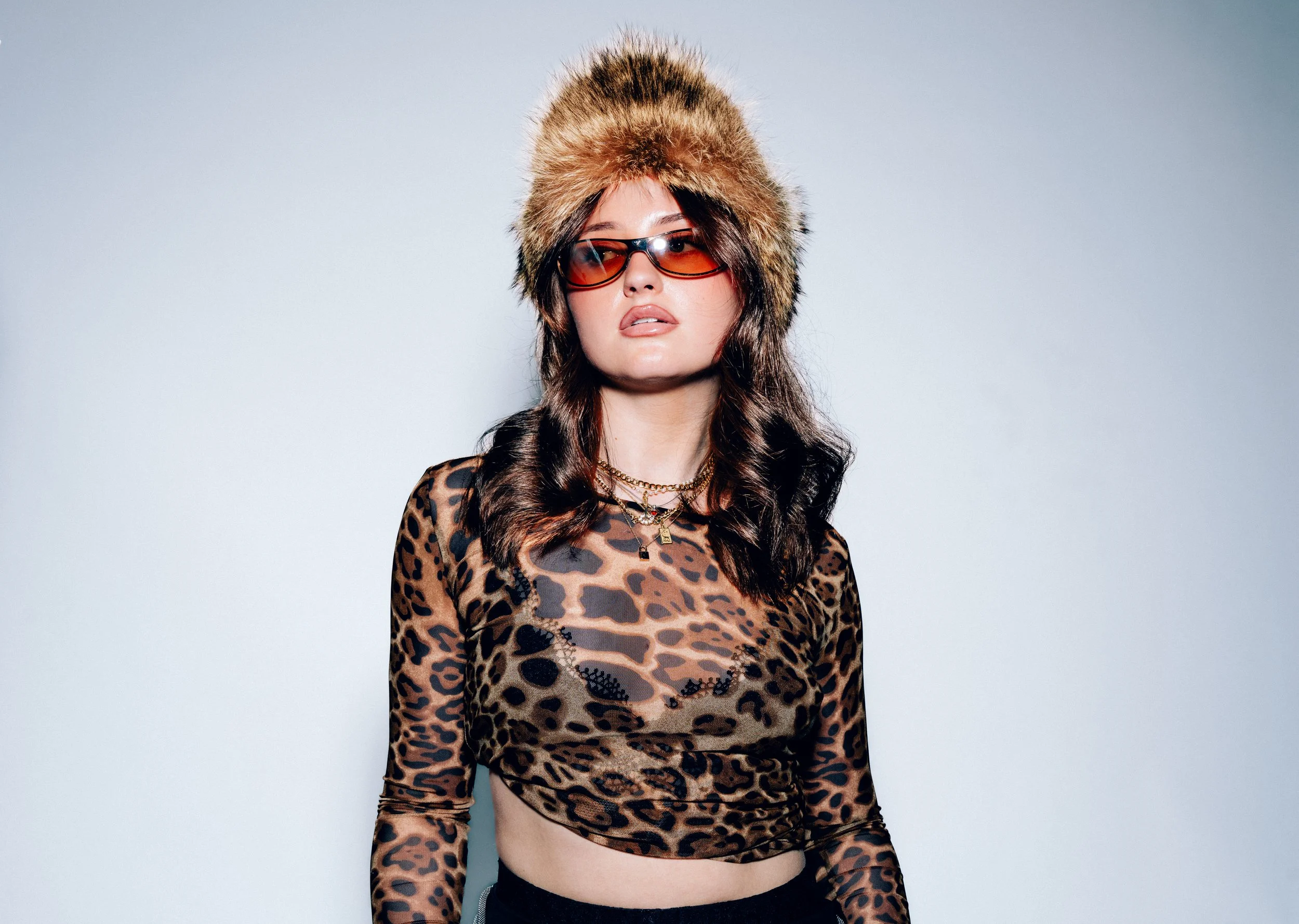 Junge Frau trägt Pelzmütze, Brille, Leopard-Muster-Oberteil und Schmuck, vor hellem Hintergrund.