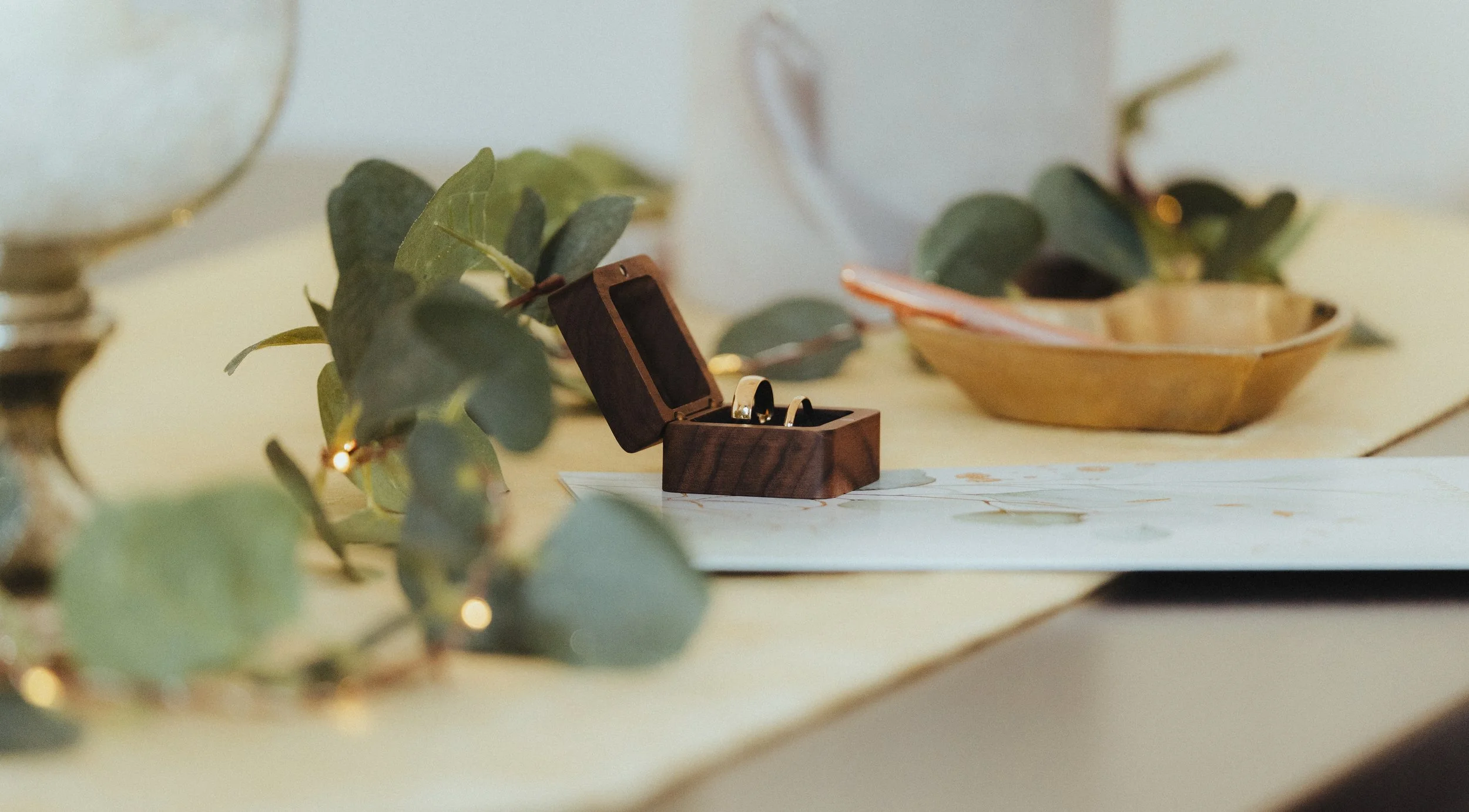 Ein offenes Holz-Schmuckkästchen mit zwei Eheringen auf einem Tisch, dekoriert mit Eukalyptuszweigen und einer goldfarbenen Schale