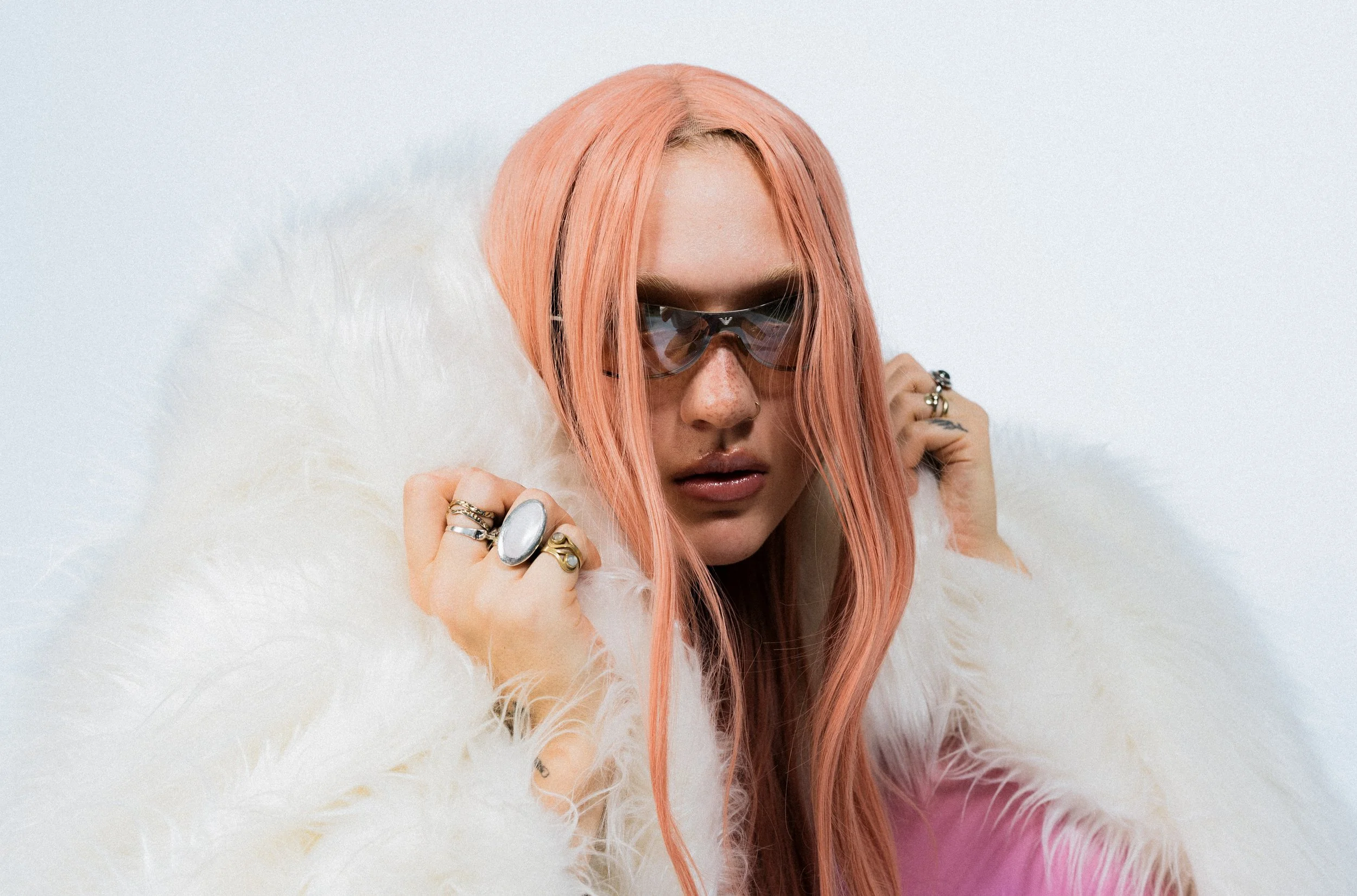 Junge Frau mit rosa Haaren, Sonnenbrille und Schmuck, umgeben von weichem, weißem Fell, gegen weißen Hintergrund.