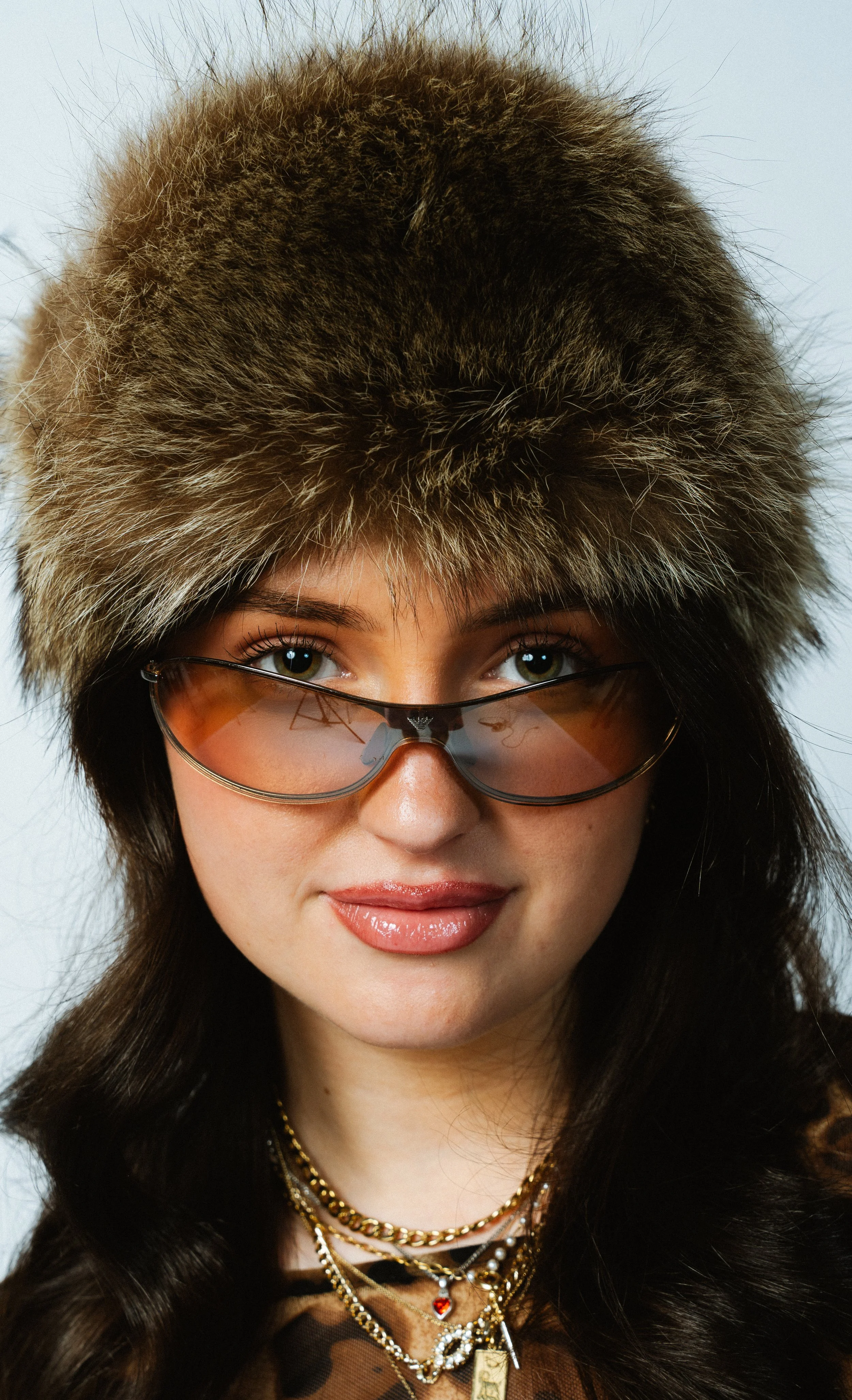Eine Frau mit langen, dunklen Haaren trägt eine große Fellmütze, orange getönte Sonnenbrille und mehrere Goldketten. Sie hat einen leichten Lächeln auf den Lippen.