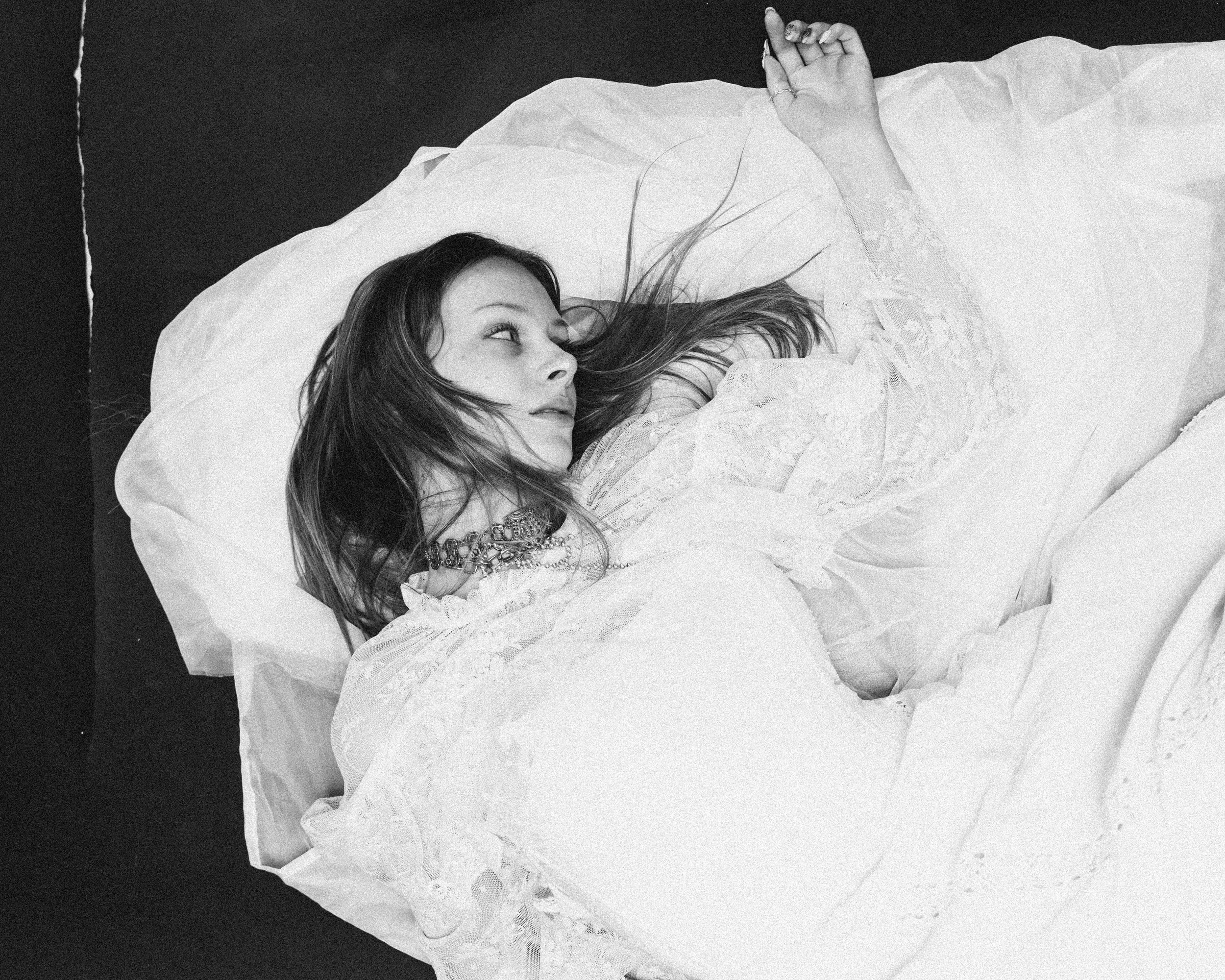 Junge Frau liegt im Bett, träumt oder schaut nach oben, trägt eine weiße, elegante Nachthemd, umgeben von Bettwäsche, schwarz-weiß Fotografie.