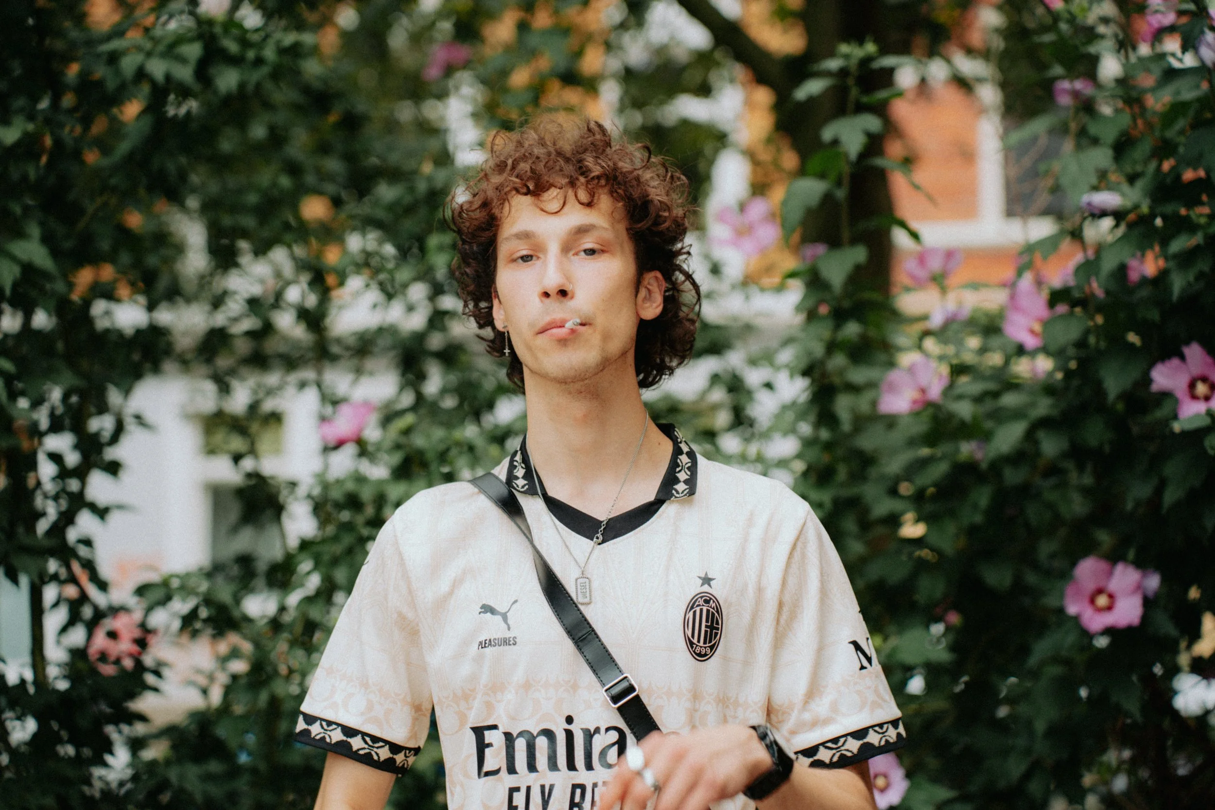 Junge mit lockigem Haar, rauchend, trägt ein AC Milan Fußballtrikot in einem Garten mit blühenden Blumen.