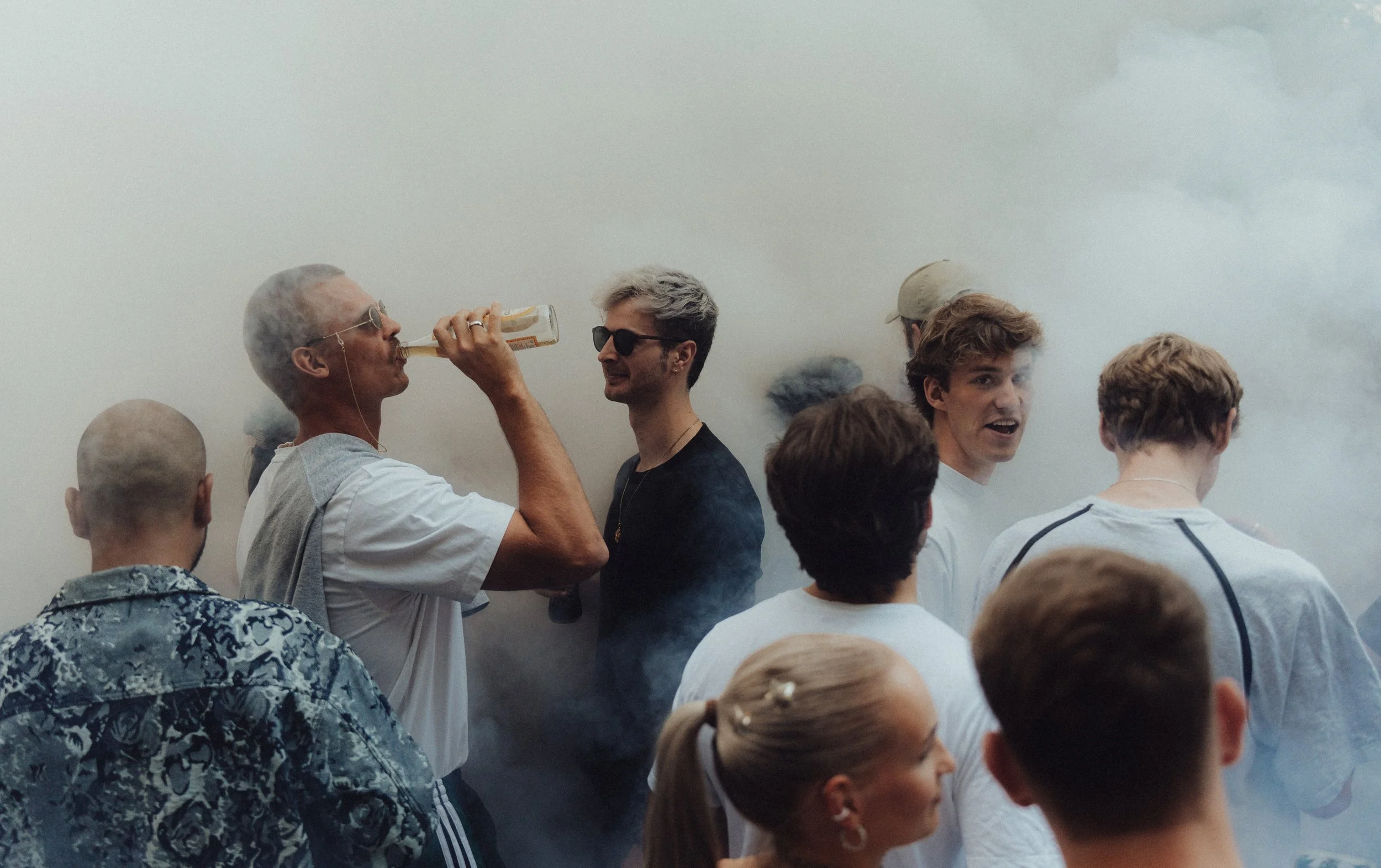 Menschen feiern in einer Gruppe, Rauch im Hintergrund, einige tragen Sonnenbrillen, eine Person trinkt ein Getränk, die Stimmung ist fröhlich.