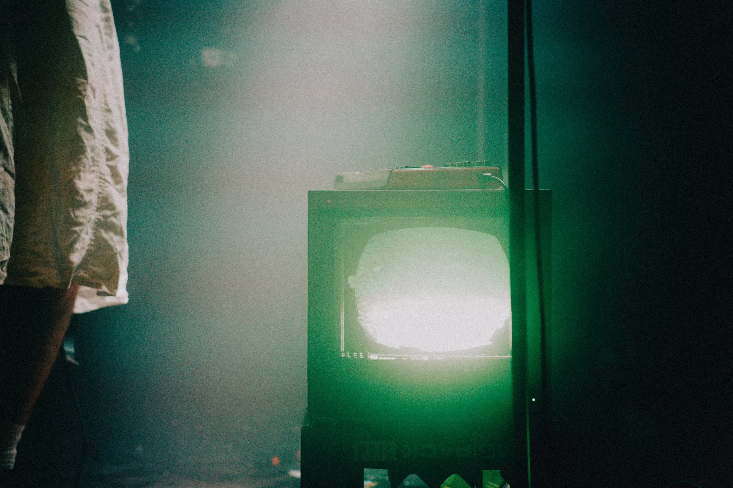 Ein alter Fernseher mit Bildstörung wird von grünem Licht beleuchtet, während eine Person im weißen Hemd im linken Bildrand nur teilweise sichtbar ist.