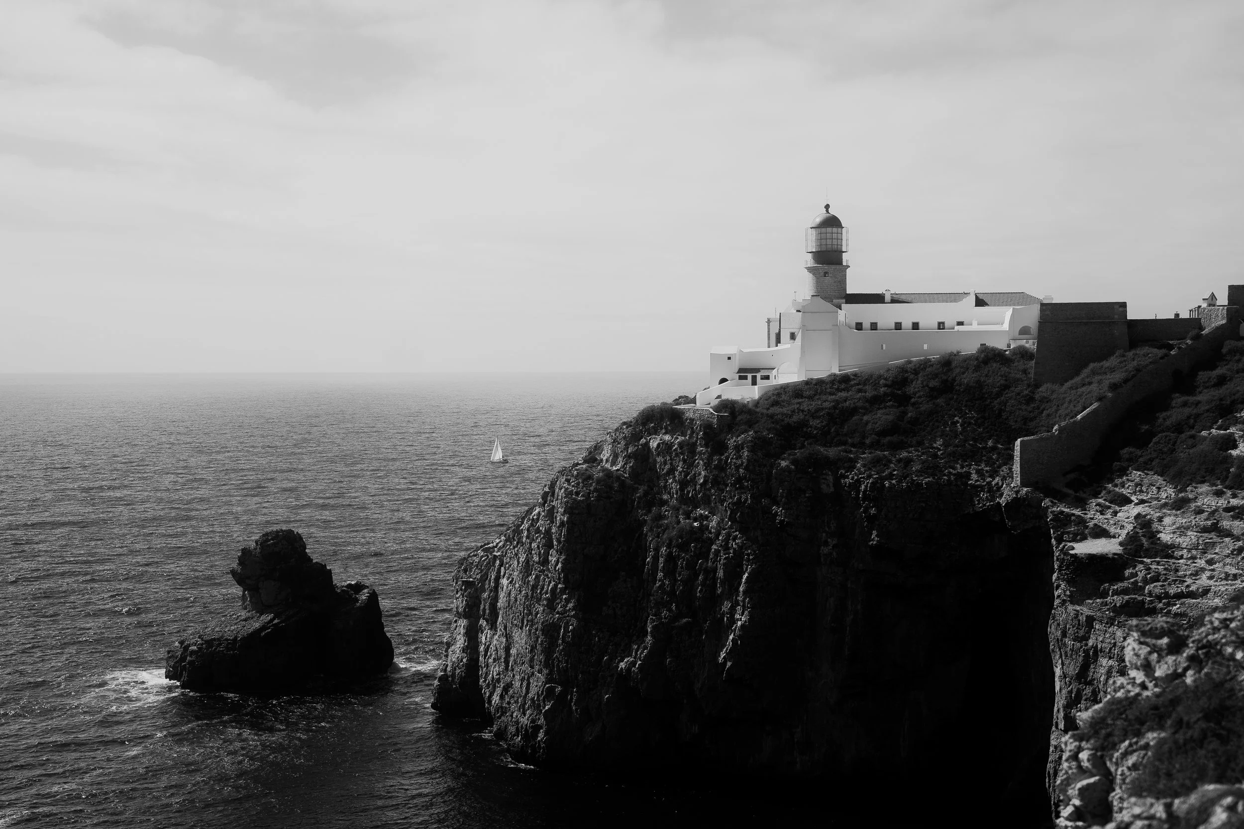 Leuchtturm auf Klippe an der Küste bei bewölktem Himmel, mit Segelboot im Meer.