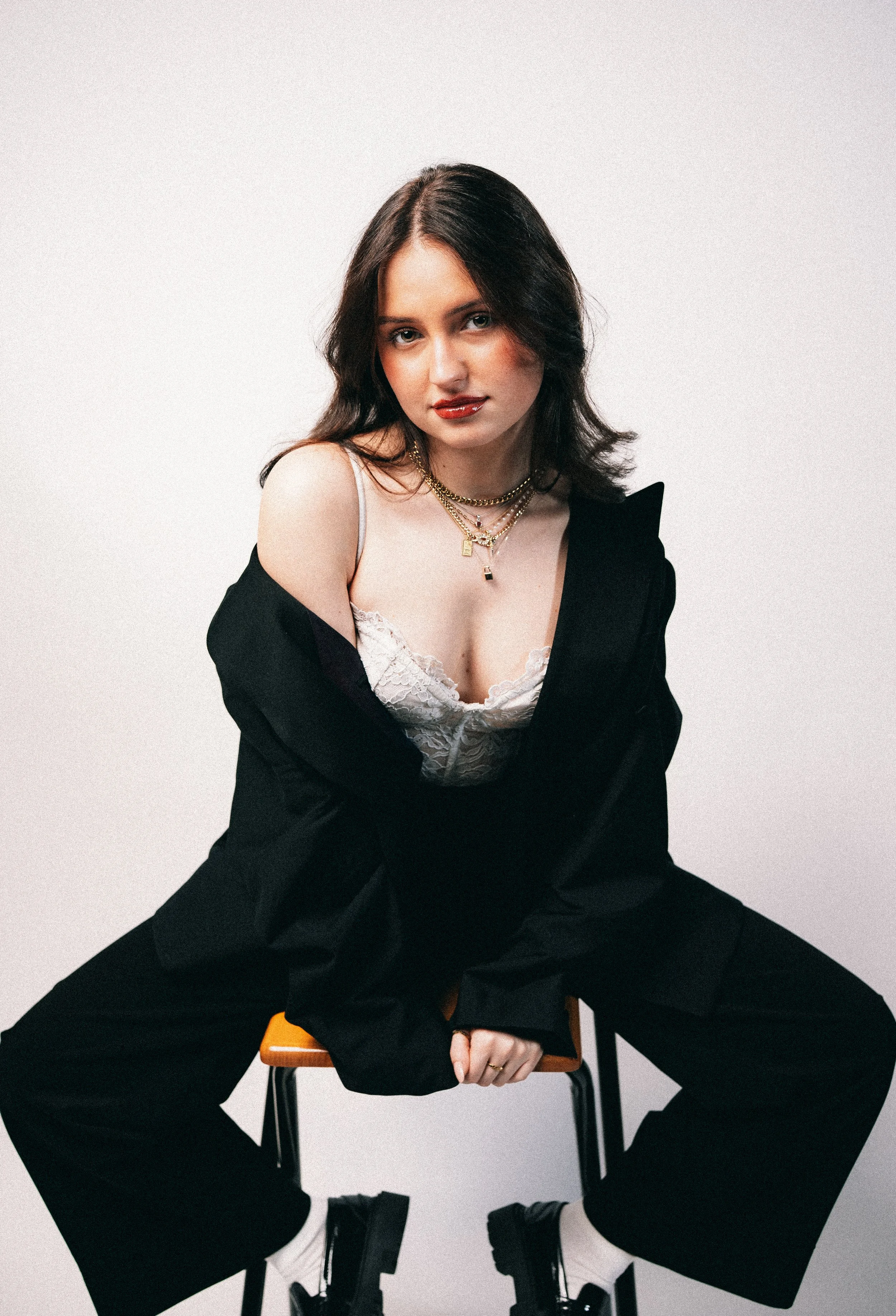Junge Frau mit dunklen Haaren, schwarzem Anzug, Spitze-Top und mehreren Halsketten sitzt auf einem Stuhl vor weißem Hintergrund.