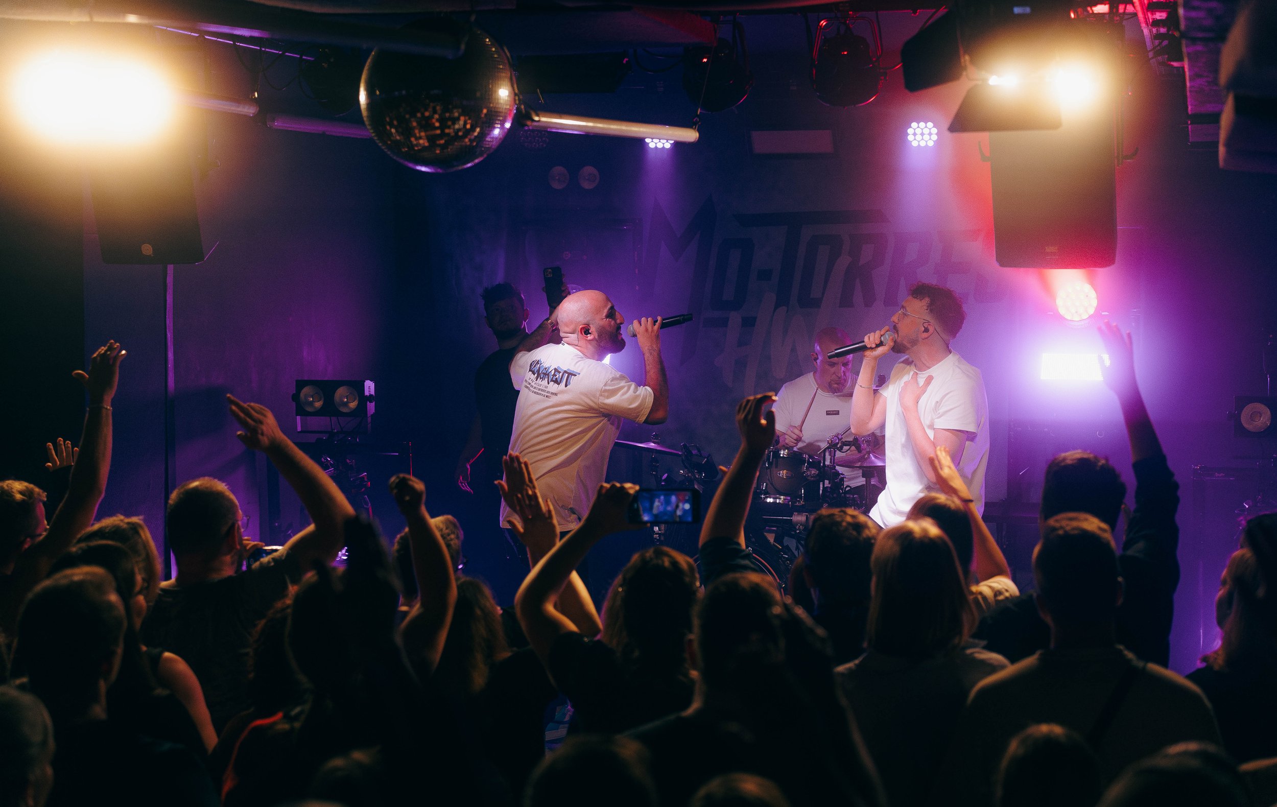 Zwei Männer singen mit Mikrophonen auf einer Bühne in einer Konzerthalle, während eine Band im Hintergrund spielt und das Publikum mit Händen in die Luft scheint.