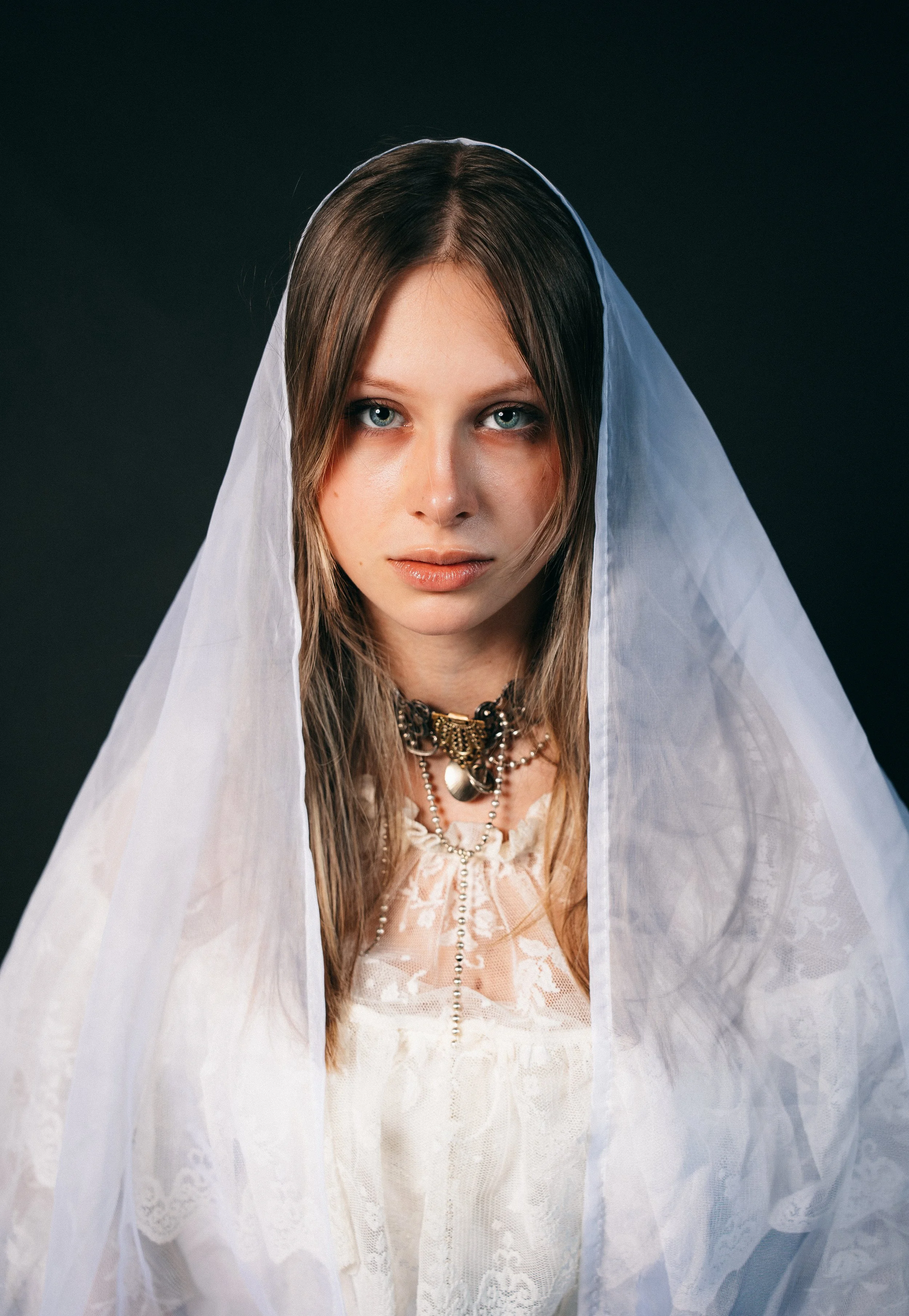 Junge Frau mit blauen Augen und langem Haar, trägt eine weiße Spitze-Tracht mit einem Schleier und mehreren Halsketten vor dunklem Hintergrund.