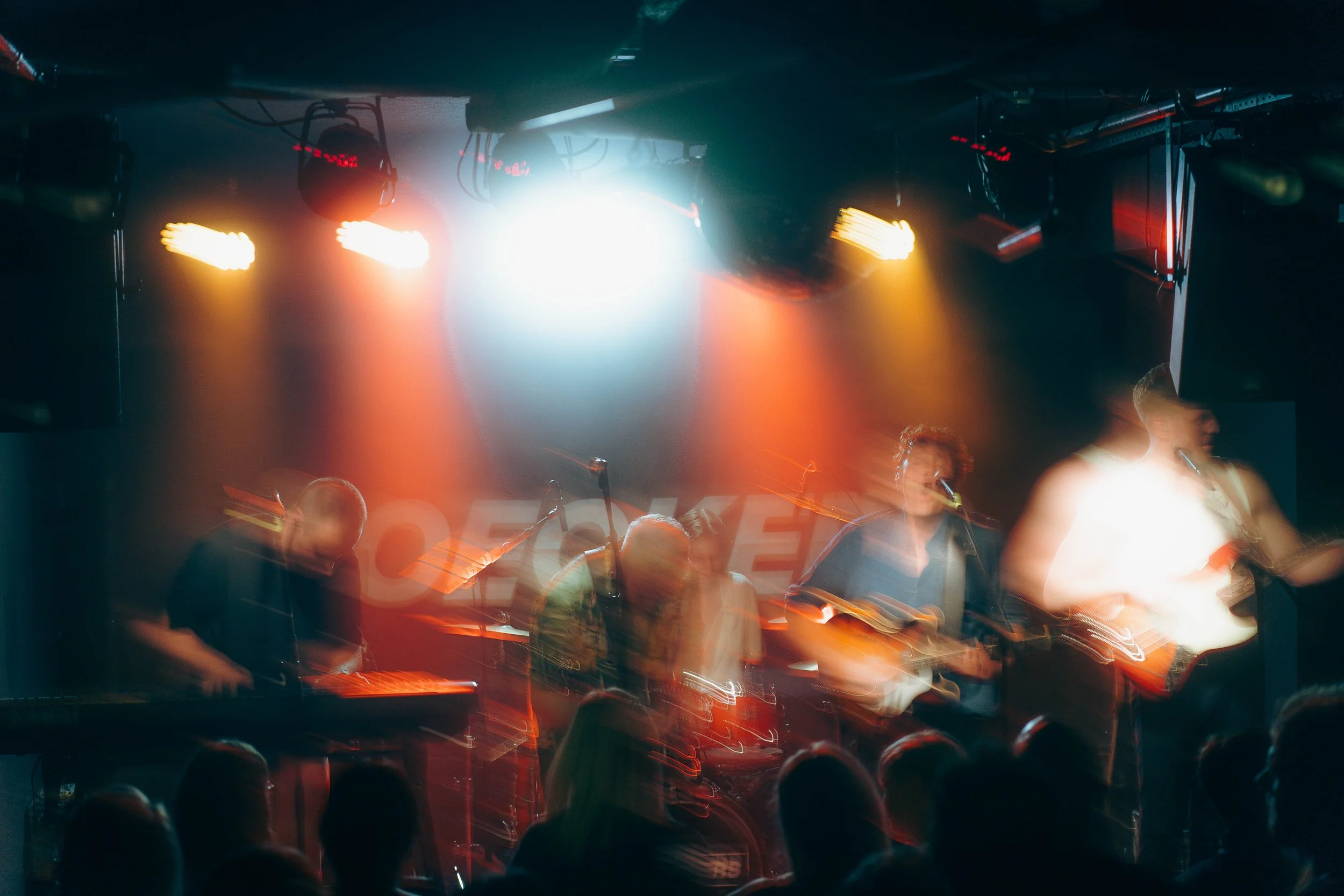 Musiker performing auf einer Bühne bei einem Konzert, mit bunten Lichtern im Hintergrund, Publikum im Vordergrund.