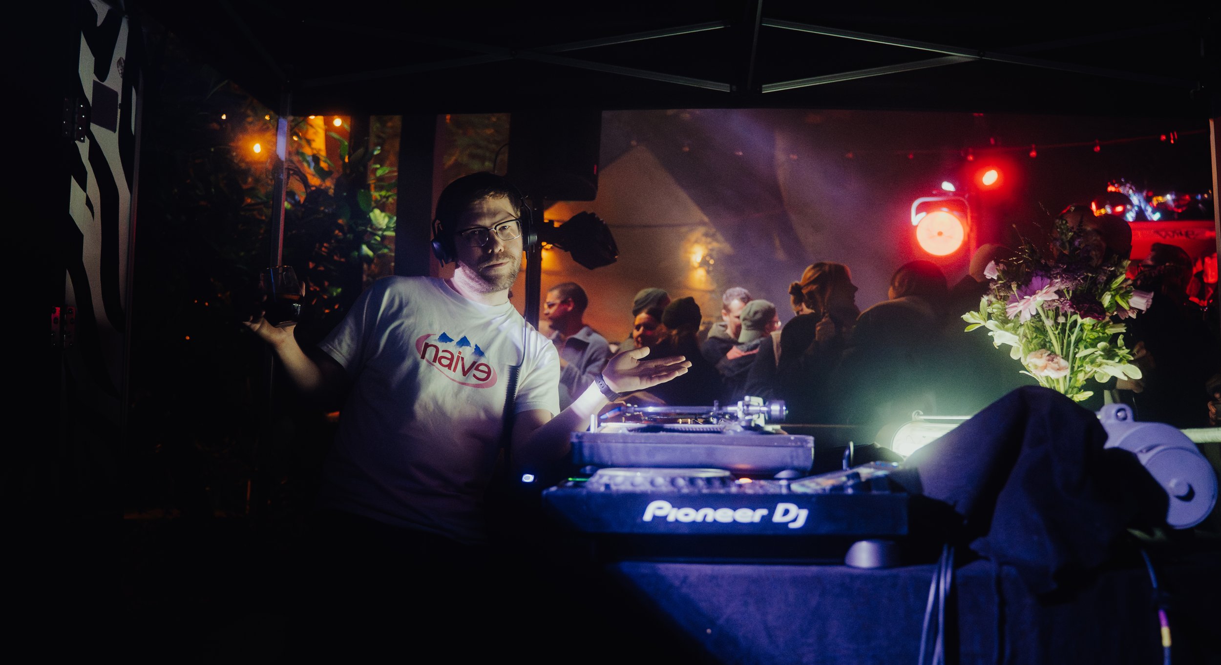 DJ mit Kopfhörern spielt in einem dunklen Club, im Hintergrund tanzen Menschen, bunte Lichteffekte und eine Blumenstrauß sind sichtbar.