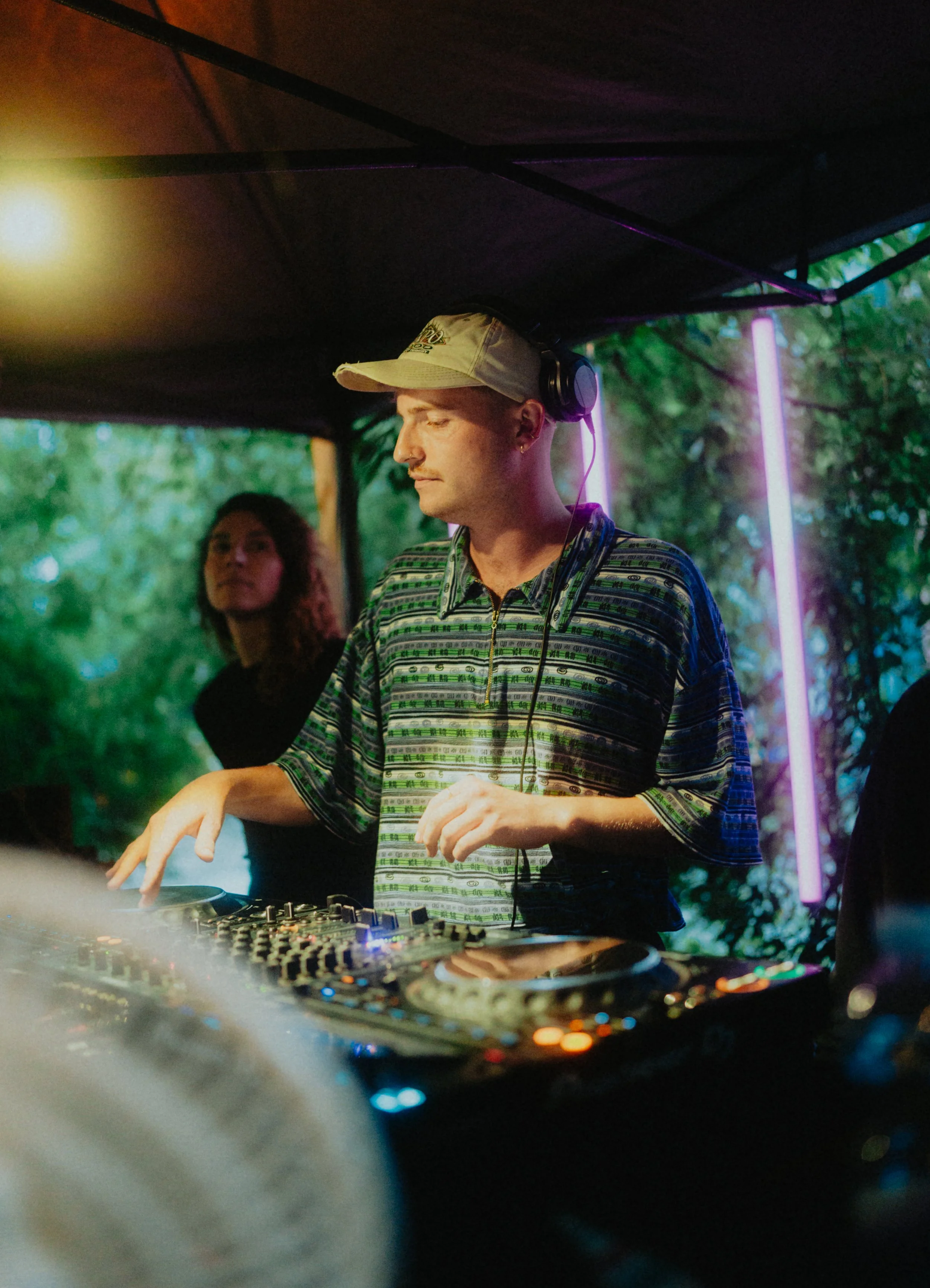 Ein DJ mit Kopfhörern und Baseballkappe spielt auf einem DJ-Controller unter einem Zelt, im Hintergrund sind Bäume und bunte Neonslichter sichtbar.