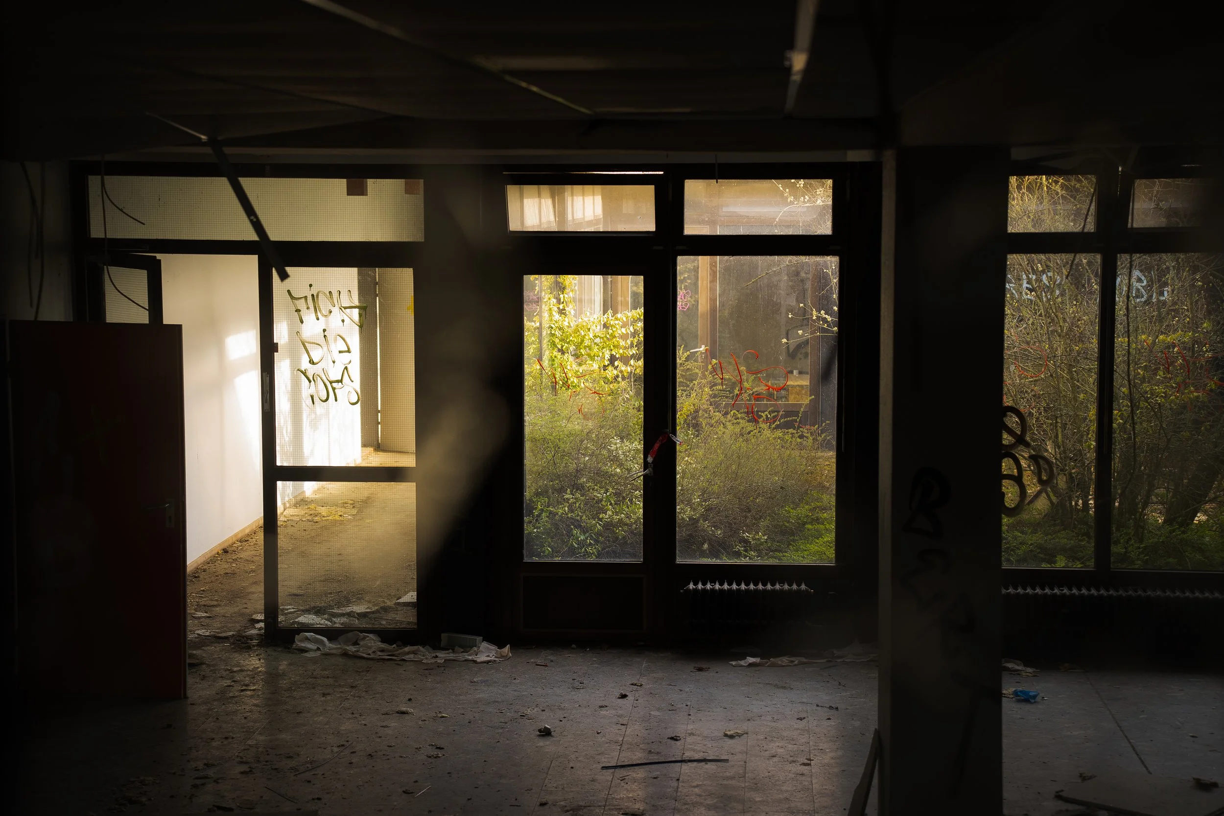 Innenansicht eines verfallenen Gebäudes mit groben Fensterfronten, Graffitis und herumliegendem Müll, im Hintergrund sichtbare grüne Bepflanzung durch die Fenster.