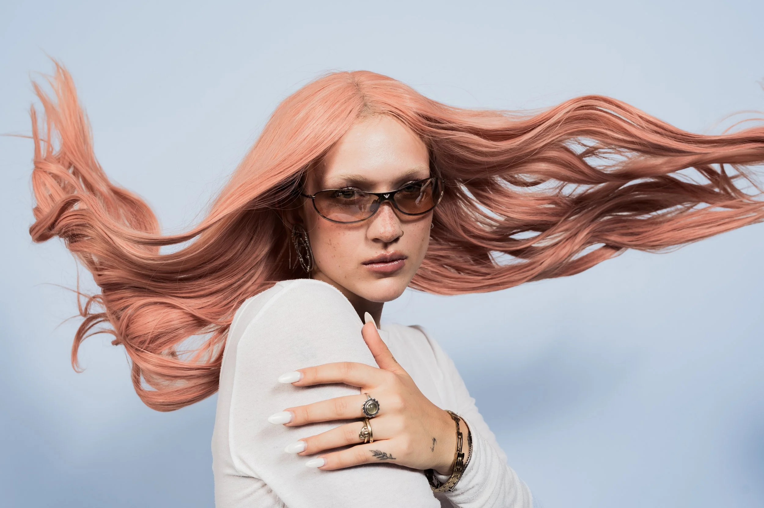 Junge Frau mit lange rosafarbenem Haar, Sonnenbrille, weißen Kleid, Ring und Armband vor hellblauem Hintergrund.