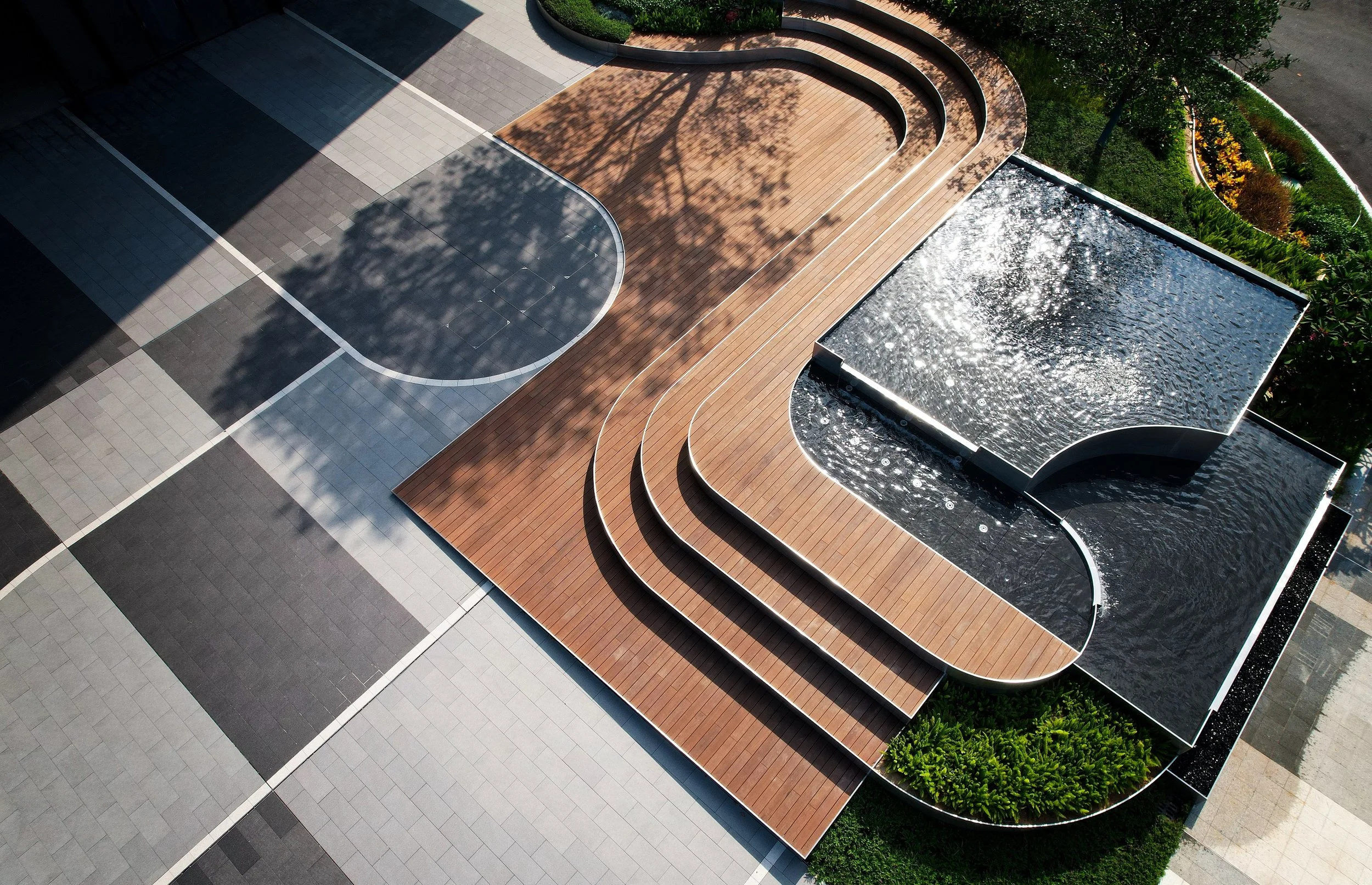 Vista aérea de un espacio público con una fuente rectangular de agua, áreas de piso de diferentes colores y un área de madera con curvas y escalones, rodeado de vegetación y árboles pequenos.