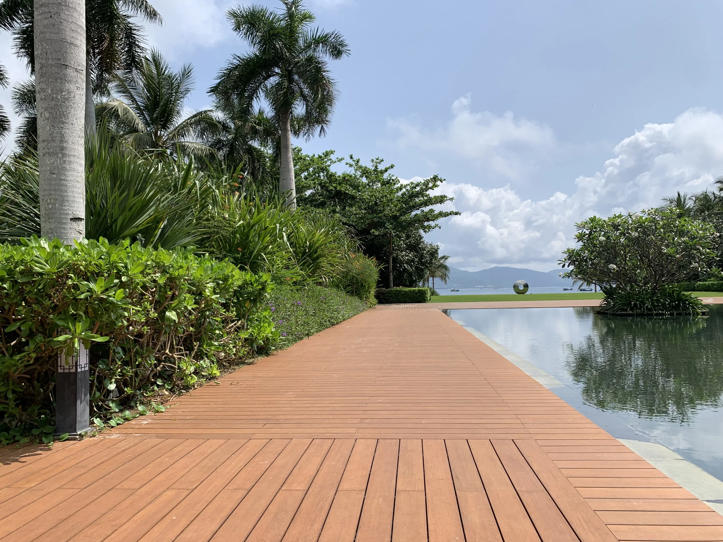 Camino de madera junto a una alberca reflejante con plantas y árboles tropicales, vista hacia el mar y montañas en el fondo, en un día soleado y con cielo parcialmente nublado.