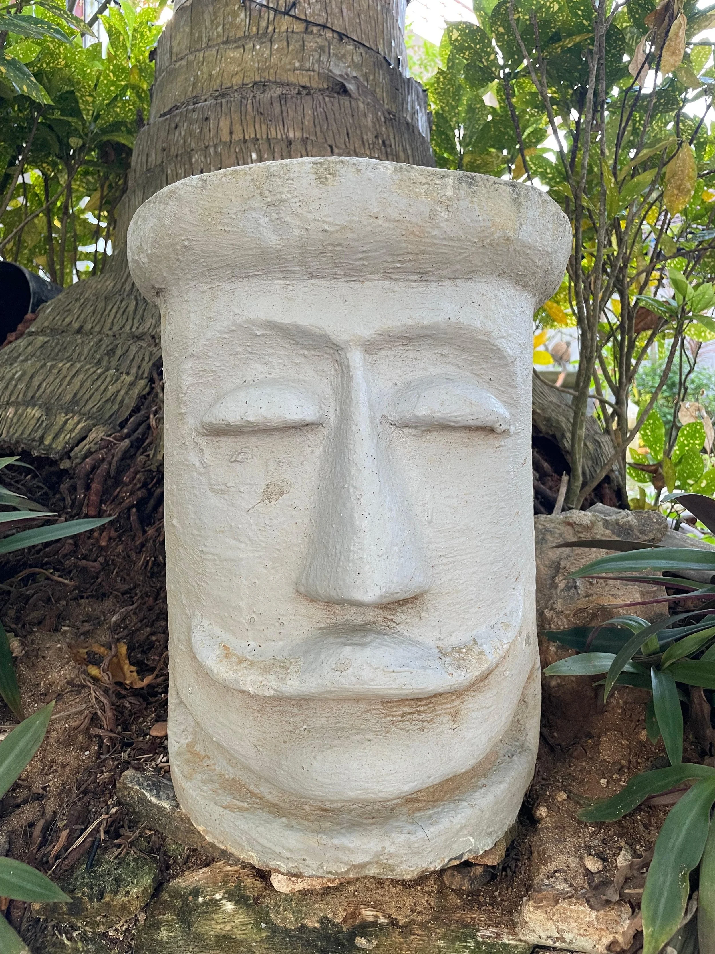 Steinerne Maske mit geschlossenen Augen, Nasenrücken, Mund und Stirn, umgeben von Pflanzen und Bäumen.