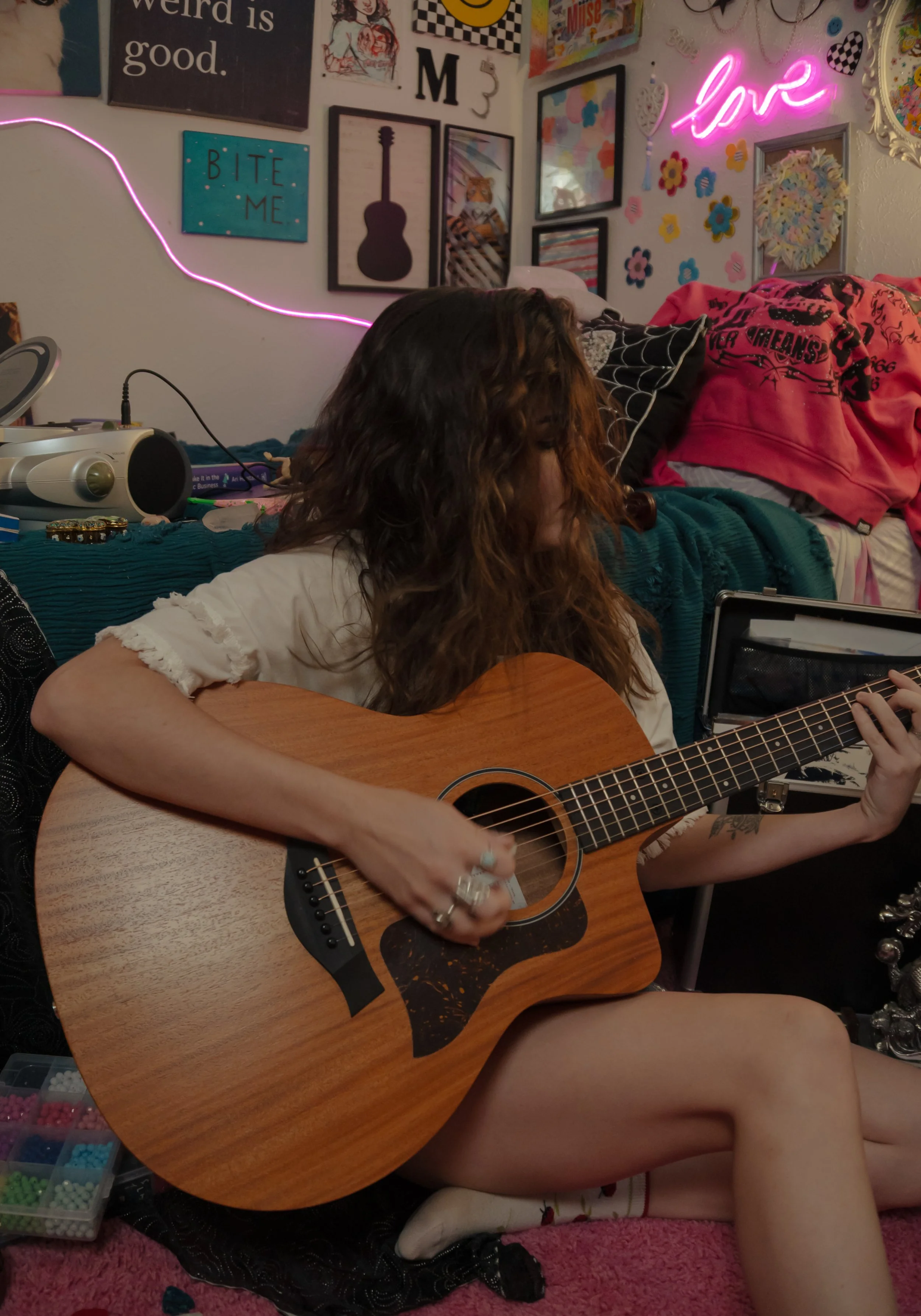 A person with long curly hair is playing an acoustic guitar in a cozy room decorated with colorful artwork, string lights, and a neon sign that says 'love'. The person is sitting on a pink carpet with their leg propped up, and a child surrounded by b