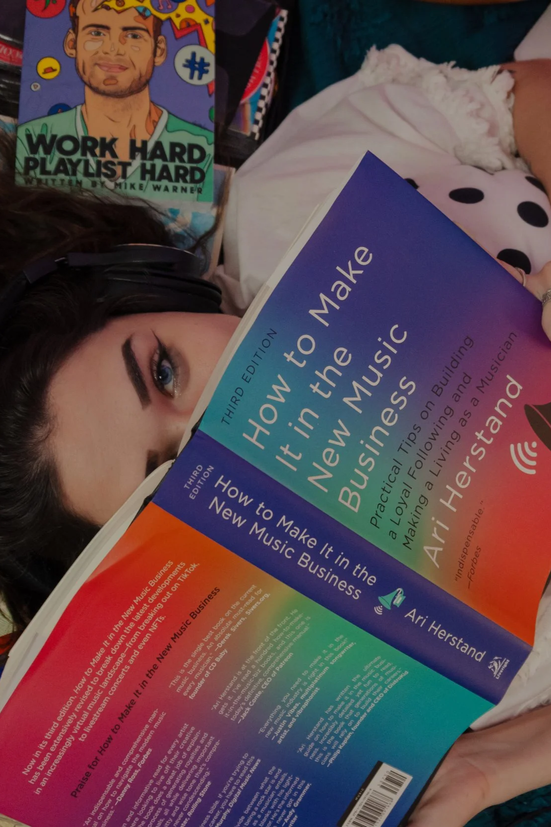 A woman with dark curly hair reading a colorful book titled 'How to Make it in the New Music Business', third edition, by Ari Herstand. The woman is wearing headphones and is partially obscured by the book.