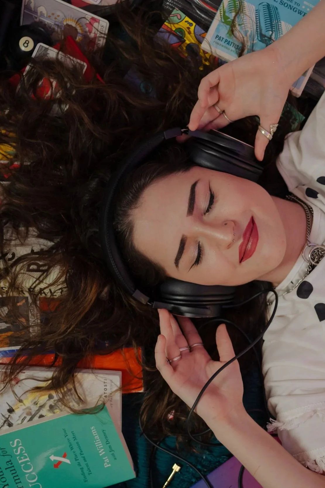 A young woman with curly dark hair, wearing headphones and makeup, lies down with her eyes closed, surrounded by books and papers.