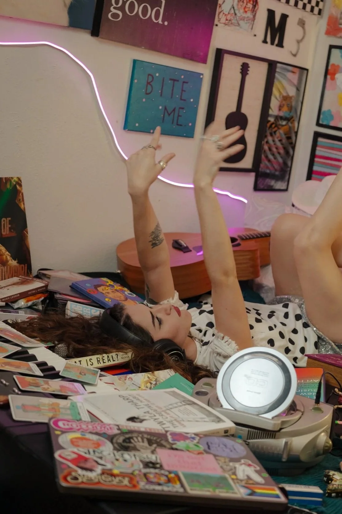 A woman lying on her back surrounded by various colorful stickers and books, wearing headphones, with arms raised, in a room decorated with artwork and a pink neon light outline.