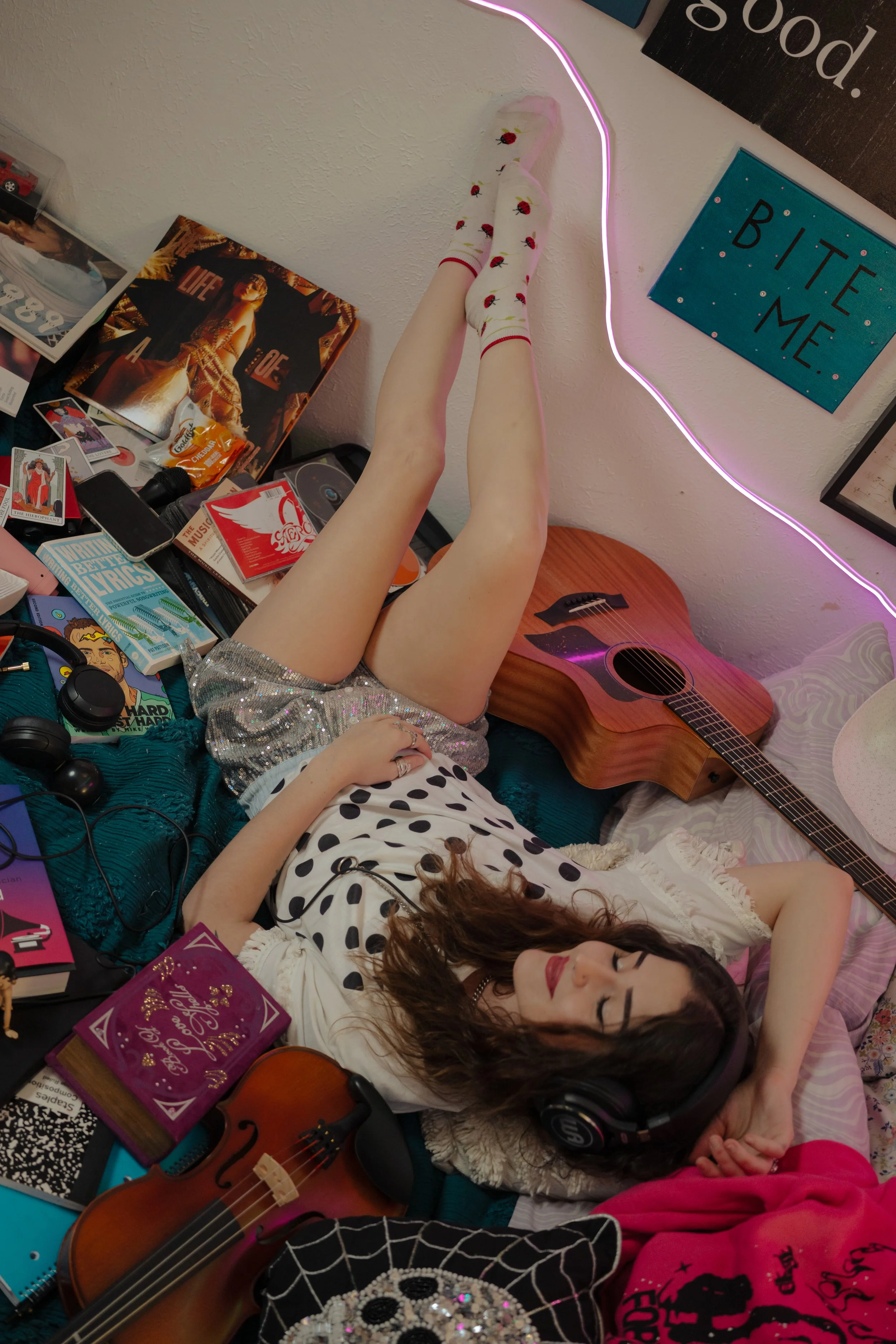 A girl with curly brown hair lying on a bed surrounded by various items including a guitar, headphones, books, and magazines, in a room decorated with posters, artwork, and neon lights.