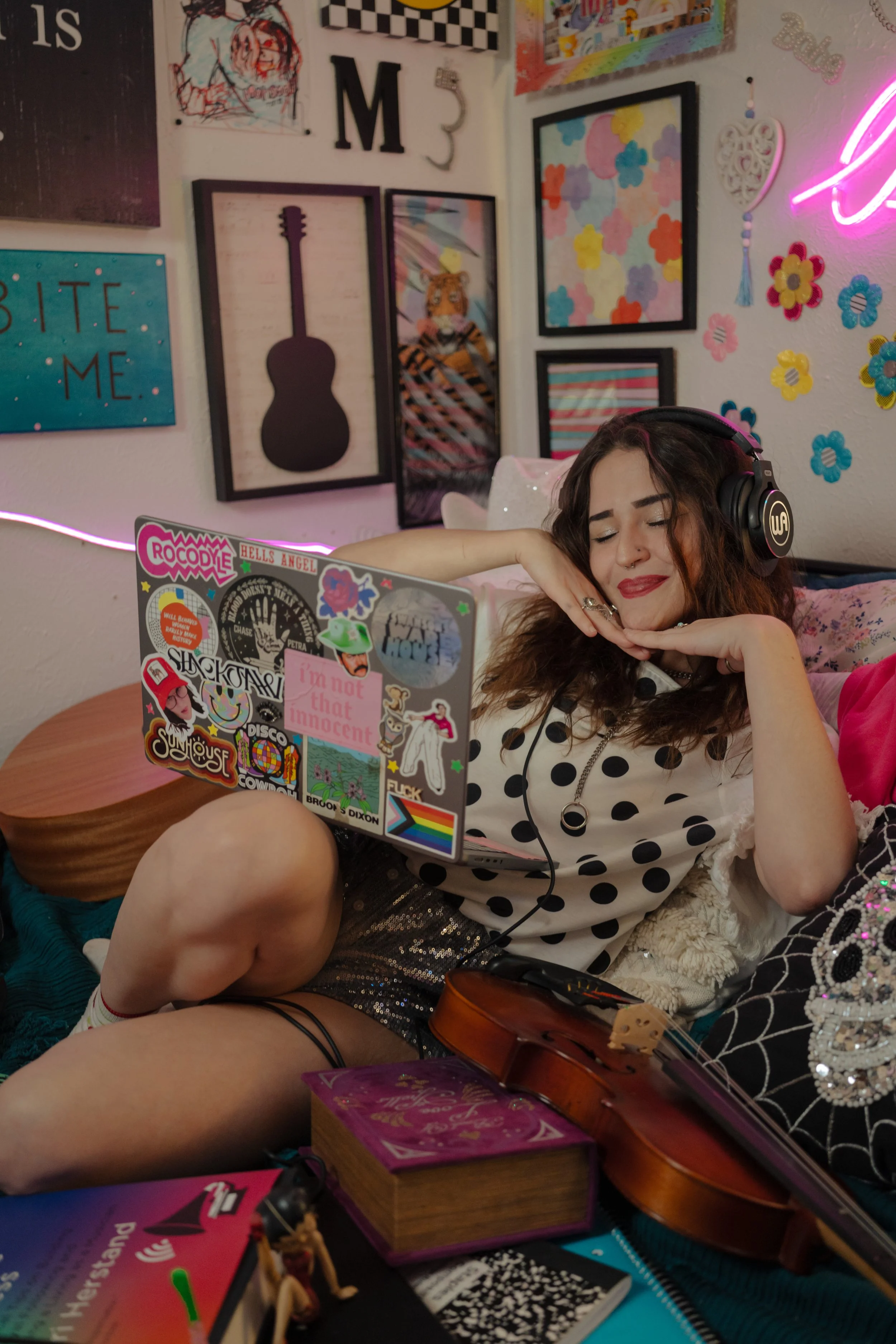 A young woman with brown hair and light skin, wearing a polka dot shirt, is sitting on the floor surrounded by musical instruments and various decorative items. She has a relaxed and happy expression, resting her head on her hands. There are posters,