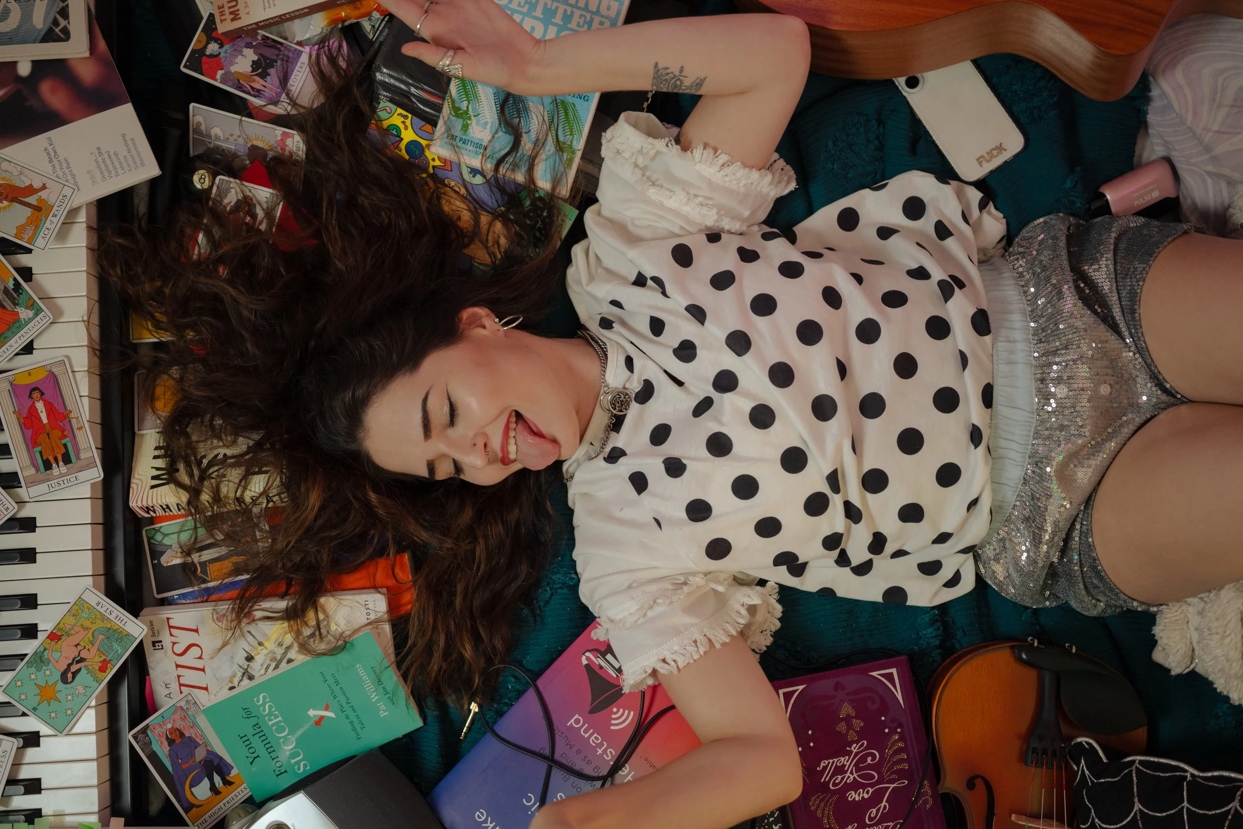 A young woman lying on a surface surrounded by tarot cards, music books, a keyboard, a smartphone, and a guitar, with her eyes closed and tongue sticking out.
