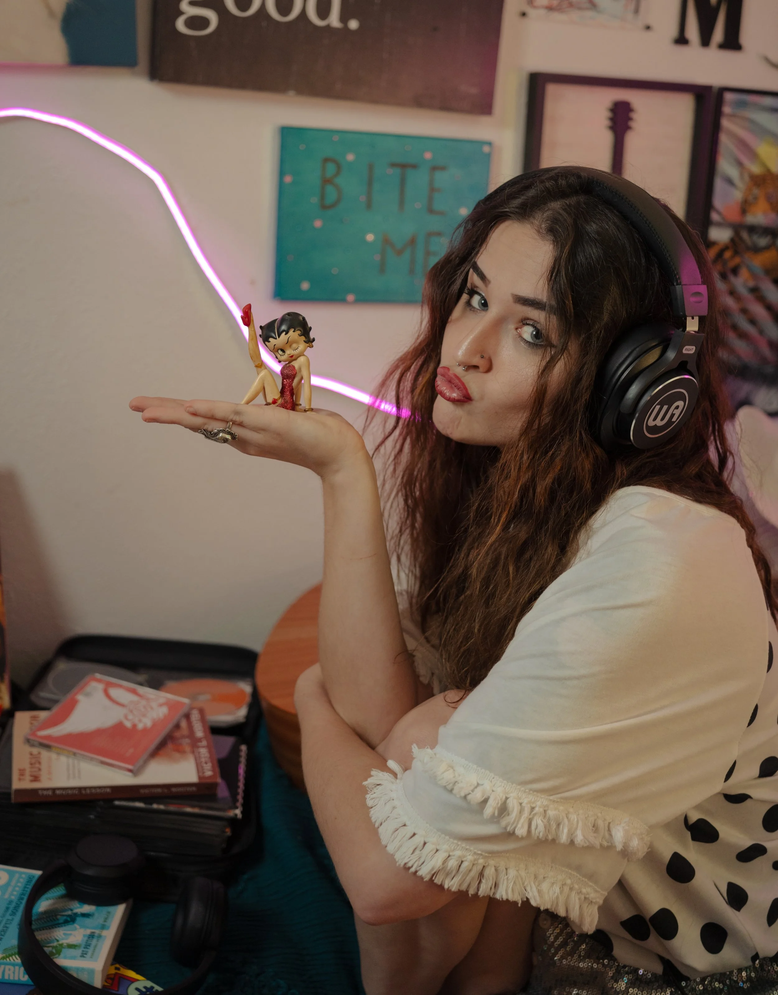 A young woman with dark hair, wearing large black headphones, holds a small Betty Boop figurine in her hand and puffs her cheeks. The background features various wall art and a table with board games and books.