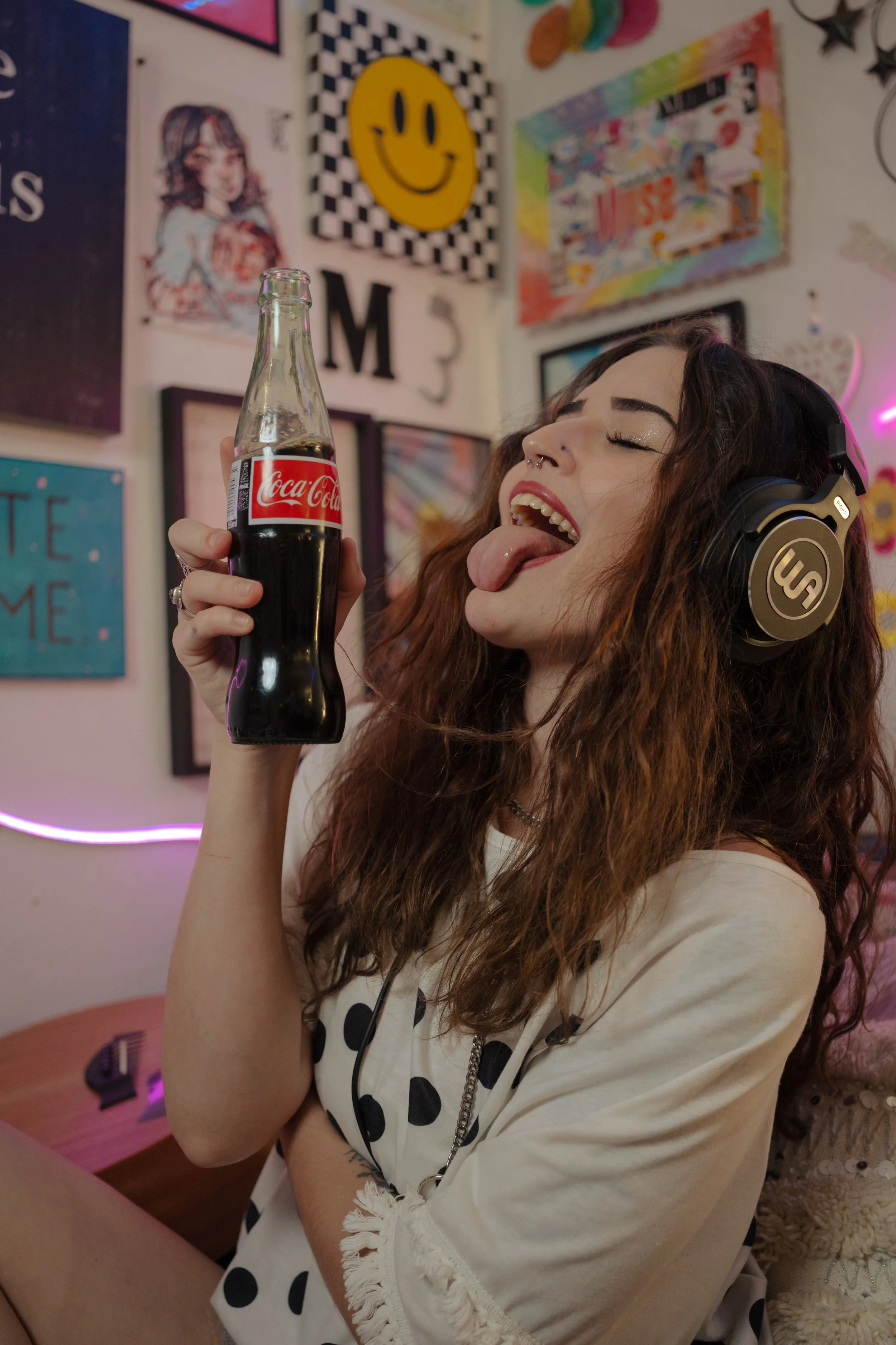 Young woman with curly hair wearing headphones with closed eyes and sticking out tongue, holding a glass Coca-Cola bottle in a colorful, decorated room.