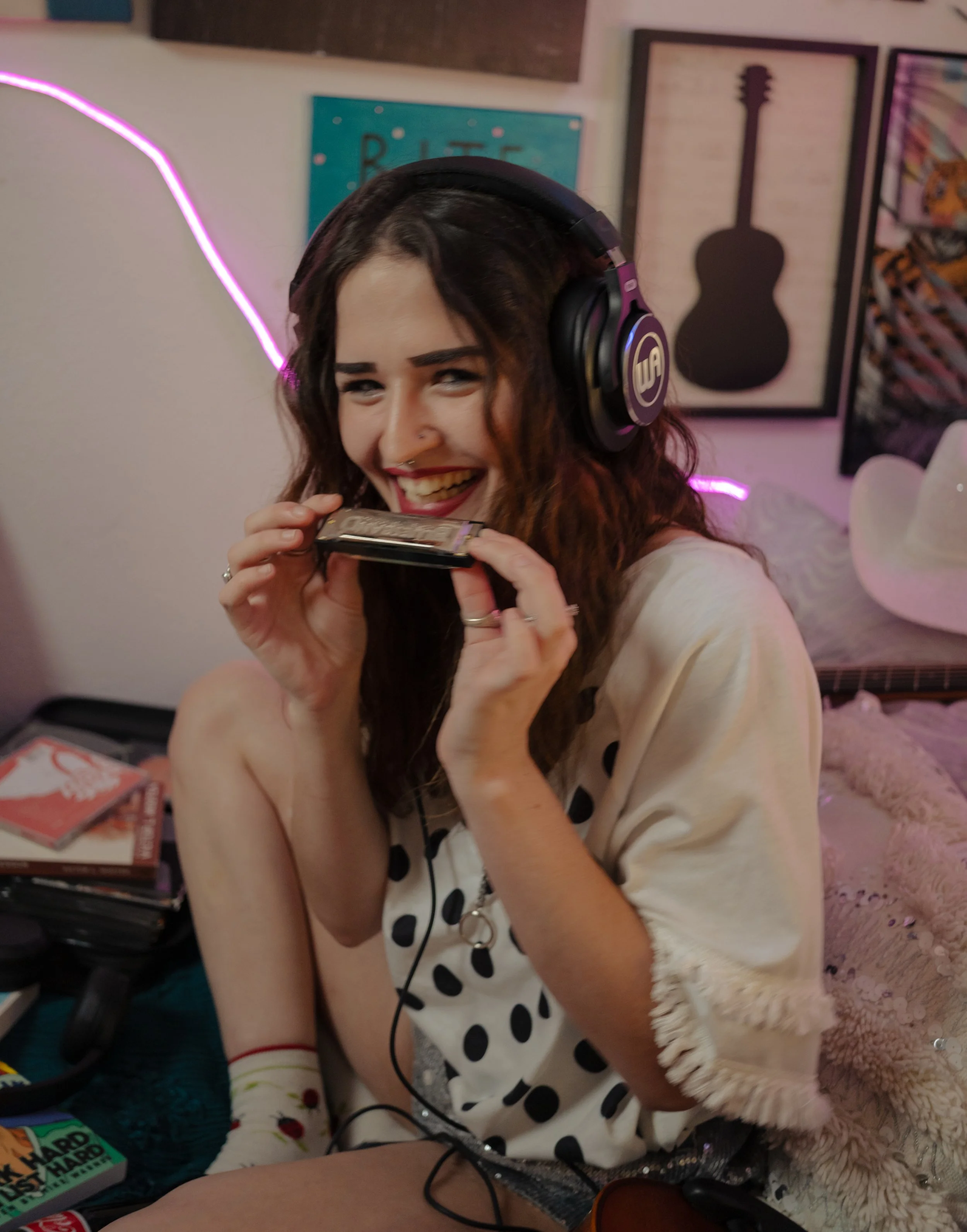 A young woman with curly brown hair, wearing headphones and a white dress with black polka dots, is smiling and holding a cassette tape. She appears to be in a cozy room with artistic posters on the wall and a pink neon light in the background.