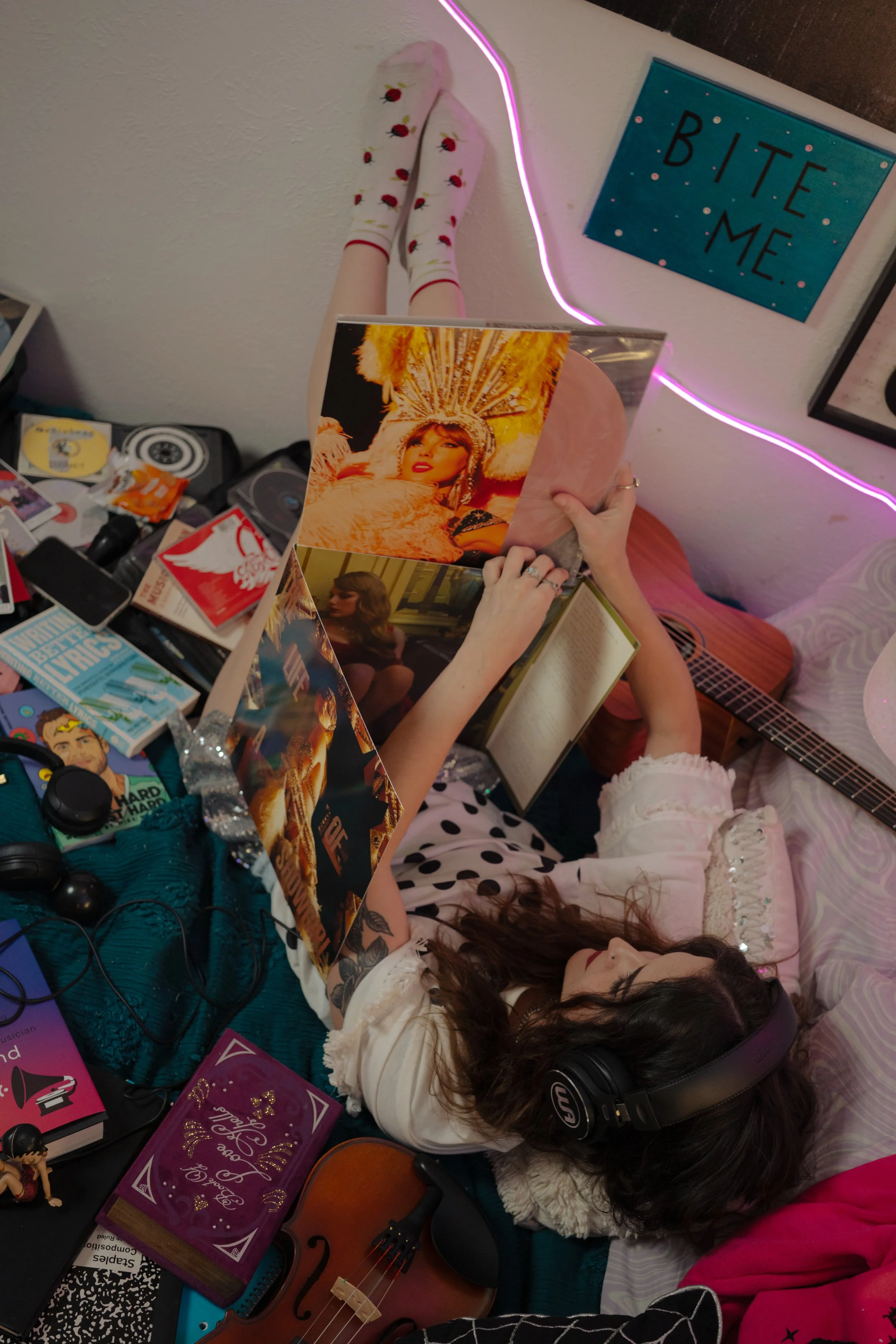 A person lying on a bed, reading a magazine with a vibrant cover featuring a woman in a headdress. The person is wearing headphones, a white top, and polka-dotted pajama pants. The room is decorated with posters, books, and a guitar. There is a pink 