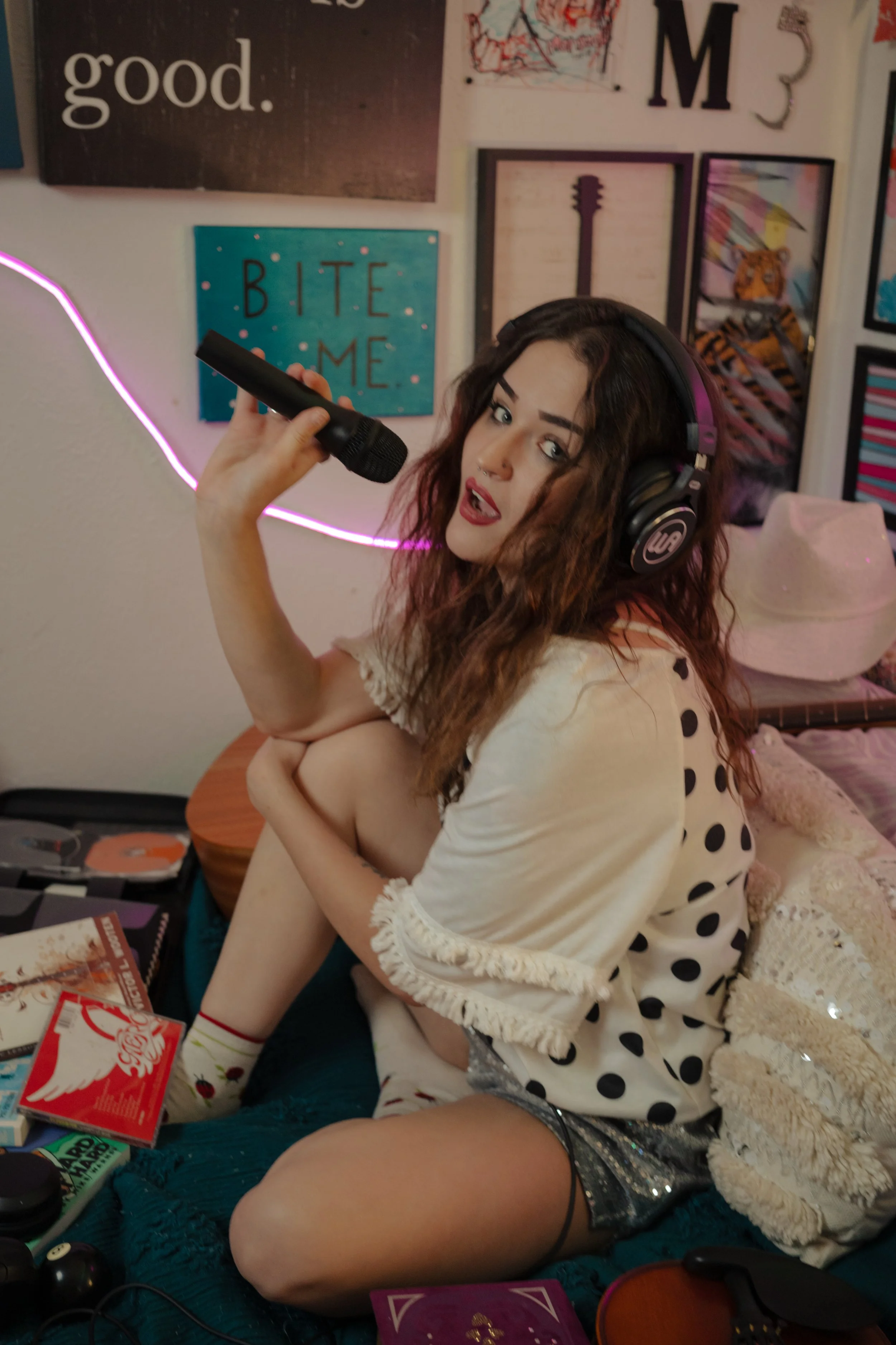 A young woman with curly hair, wearing headphones and a polka dot shirt, holding a microphone and posing indoors in front of wall art.