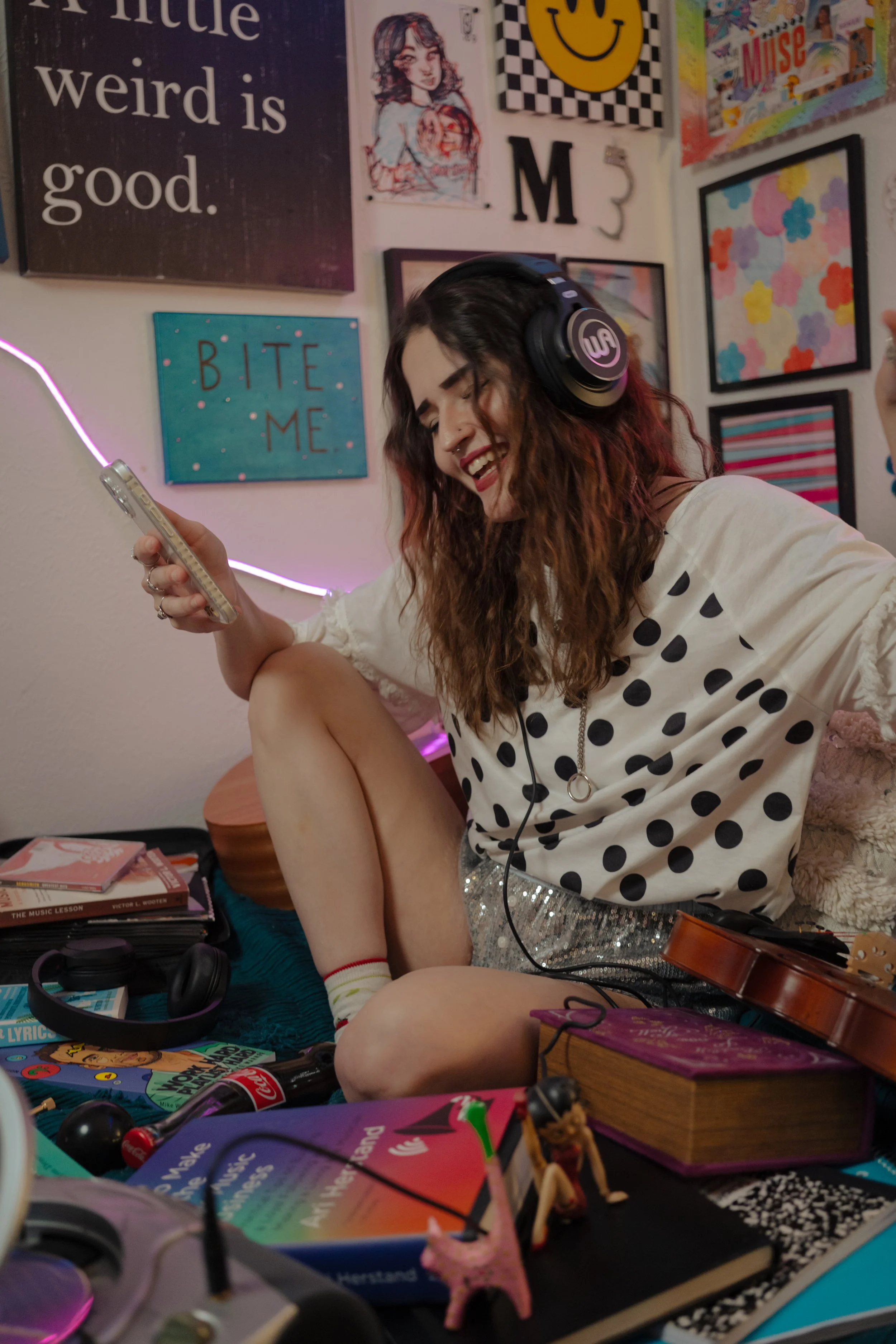 A young woman with wavy brown hair wearing headphones, a white polka dot shirt, and glittery shorts, sitting on the floor and smiling while looking at her phone with a colorful, eclectic wall art background. The wall art includes a smiley face, a gir