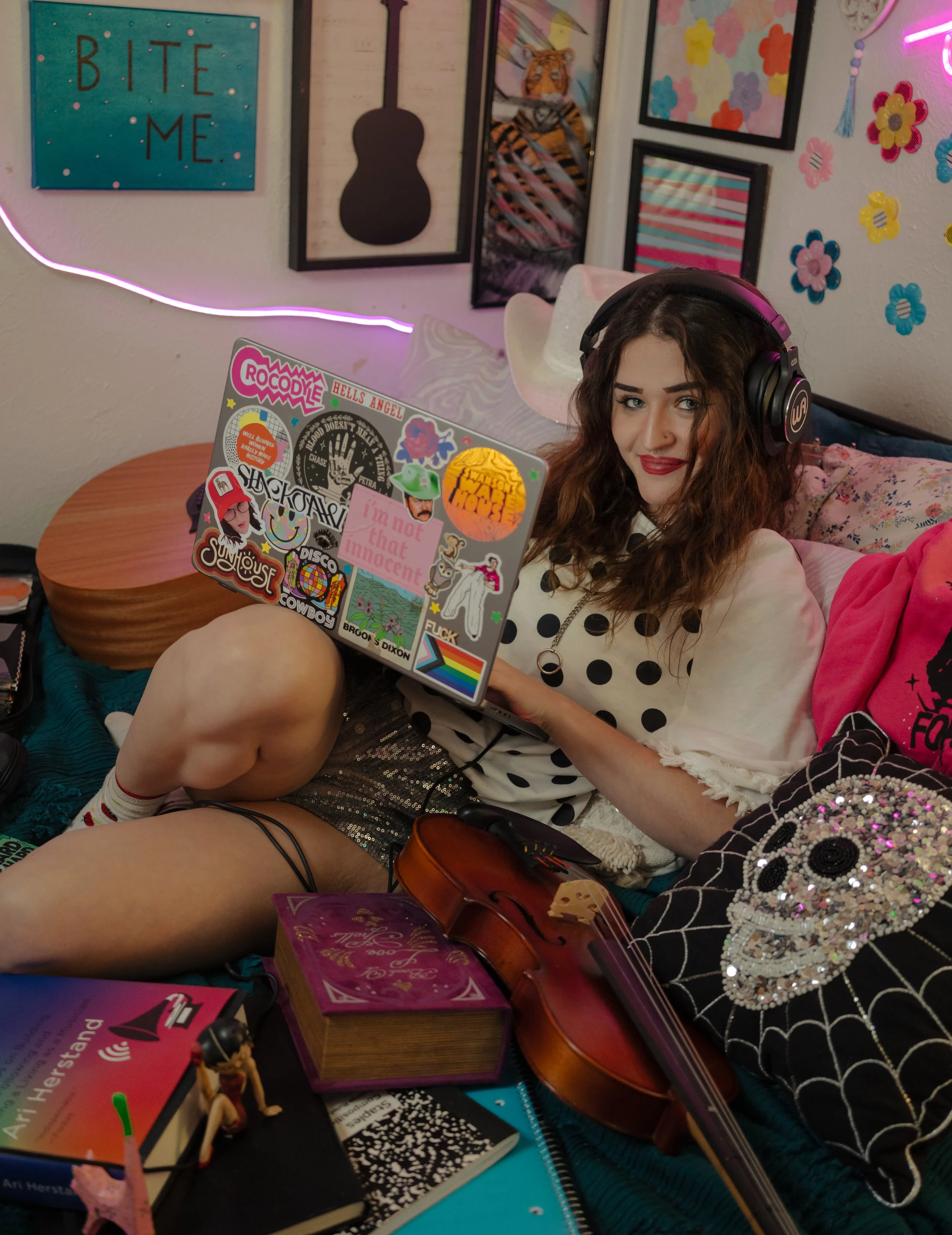 A young woman with curly brown hair, wearing headphones and a white shirt with black polka dots, is sitting on a bed in a decorated room. She is smiling and holding a laptop covered with various stickers. The room features colorful wall art, includin
