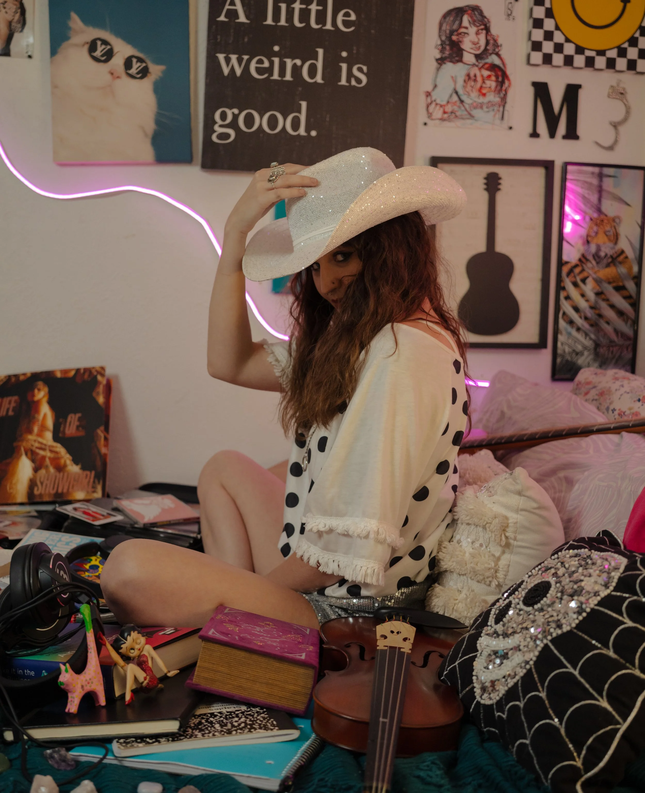 A woman with curly hair wearing a white polka-dotted top and a glittery cowboy hat, sitting amidst a cluttered room with various posters and art on the walls, and objects on the table including plush toys, books, and decorative items.
