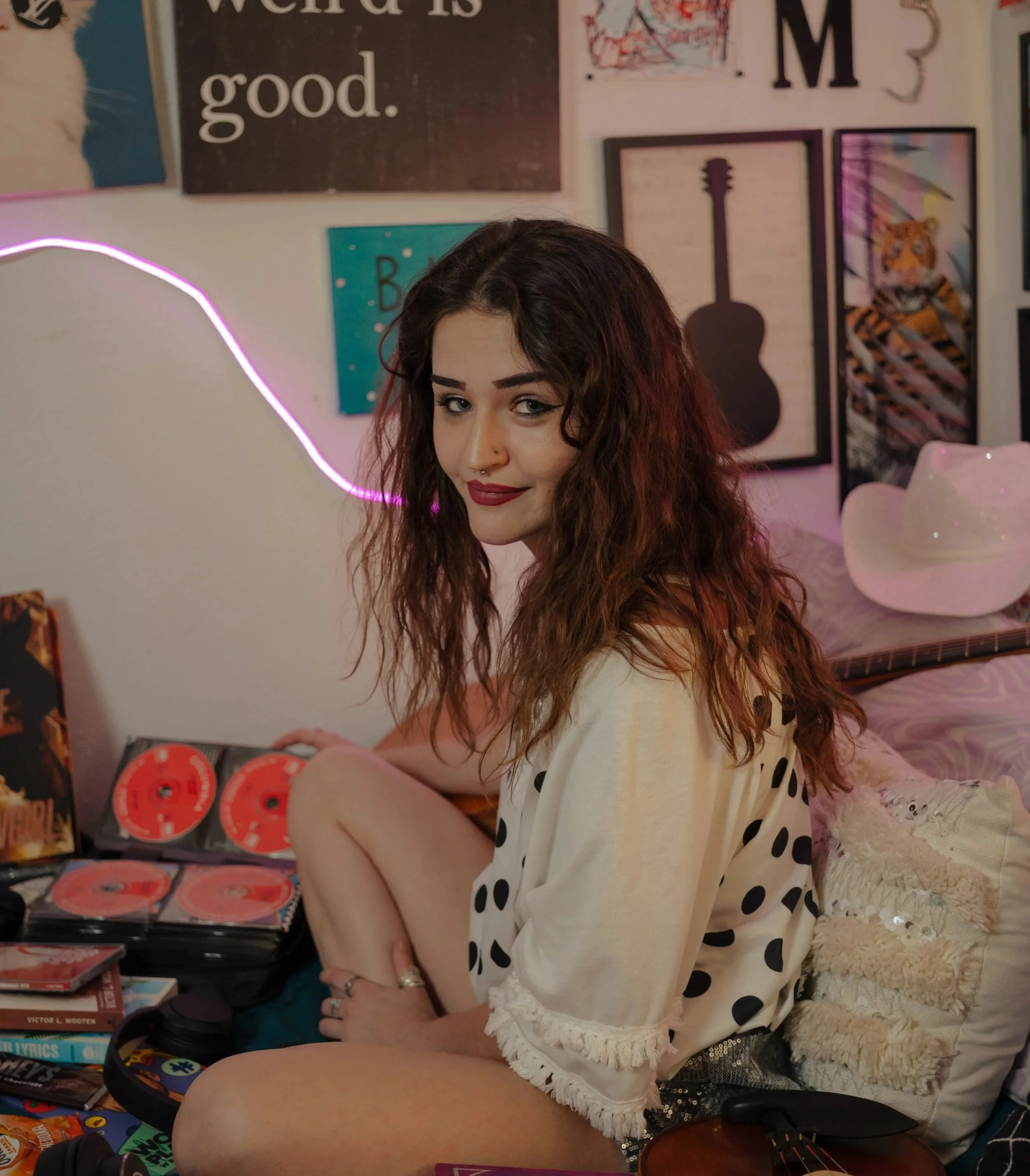 A young woman with curly brown hair, wearing a white polka-dotted shirt with lace trim, sitting in front of a wall decorated with posters, one with the word 'good', another with a guitar silhouette, and others featuring abstract designs and animals. She holds a multicolored item in her hands and has a slight smile on her face.