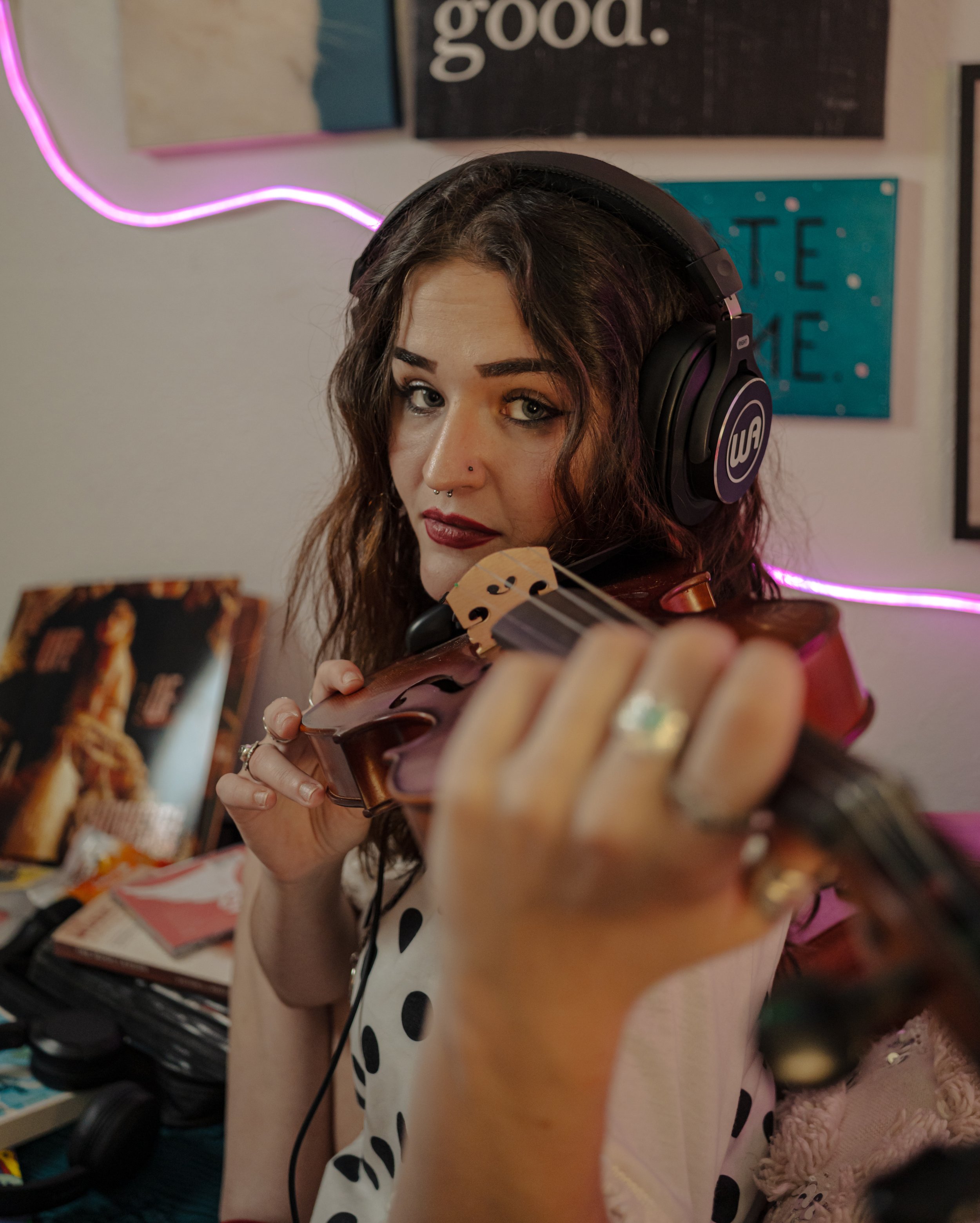 A woman with brown hair and tattoos on her arm holds a violin and a bow in front of a wall decorated with posters and neon lights, with a blurred person in the background.