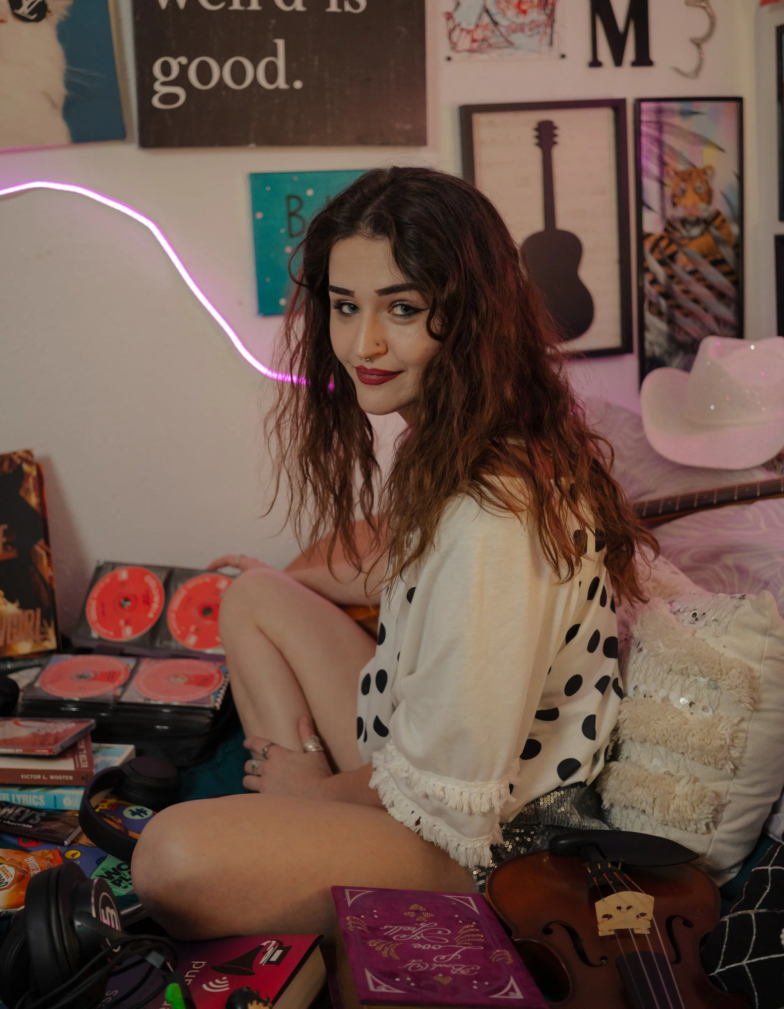 A young woman with dark curly hair and makeup wearing a cream-colored top with black polka dots sitting at a desk covered with books, a purple notebook, a pair of headphones, and a guitar. The background features a wall decorated with art, posters, a