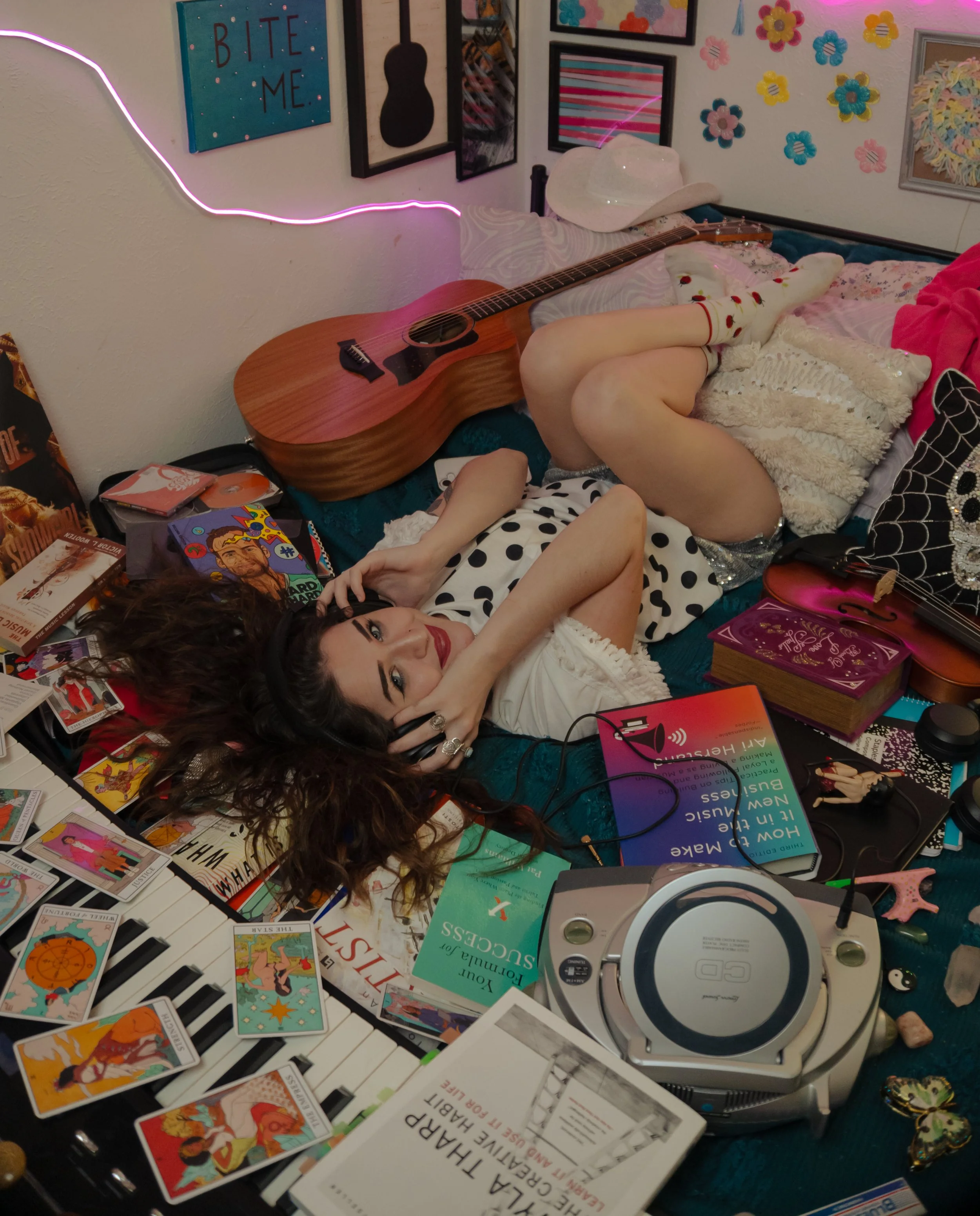 A young woman with dark, curly hair and red lipstick lies on a bed surrounded by various books, magazines, and tarot cards. She is wearing a white shirt with black polka dots and is holding headphones. The wall behind her features colorful artwork, i