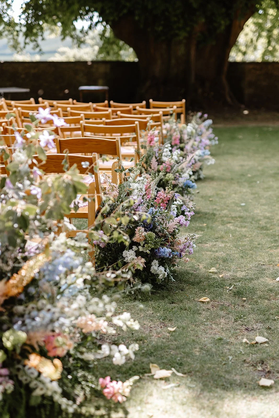 Floral meadow arrangements lining the ceremony aisle beneath the ancient lime tree at Hamswell House in Bath, designed for a June wedding. Garden-style flowers included Astilbe, Stocks, Delphinium, Eremurus (foxtail lilies), Clematis and Nigella in a