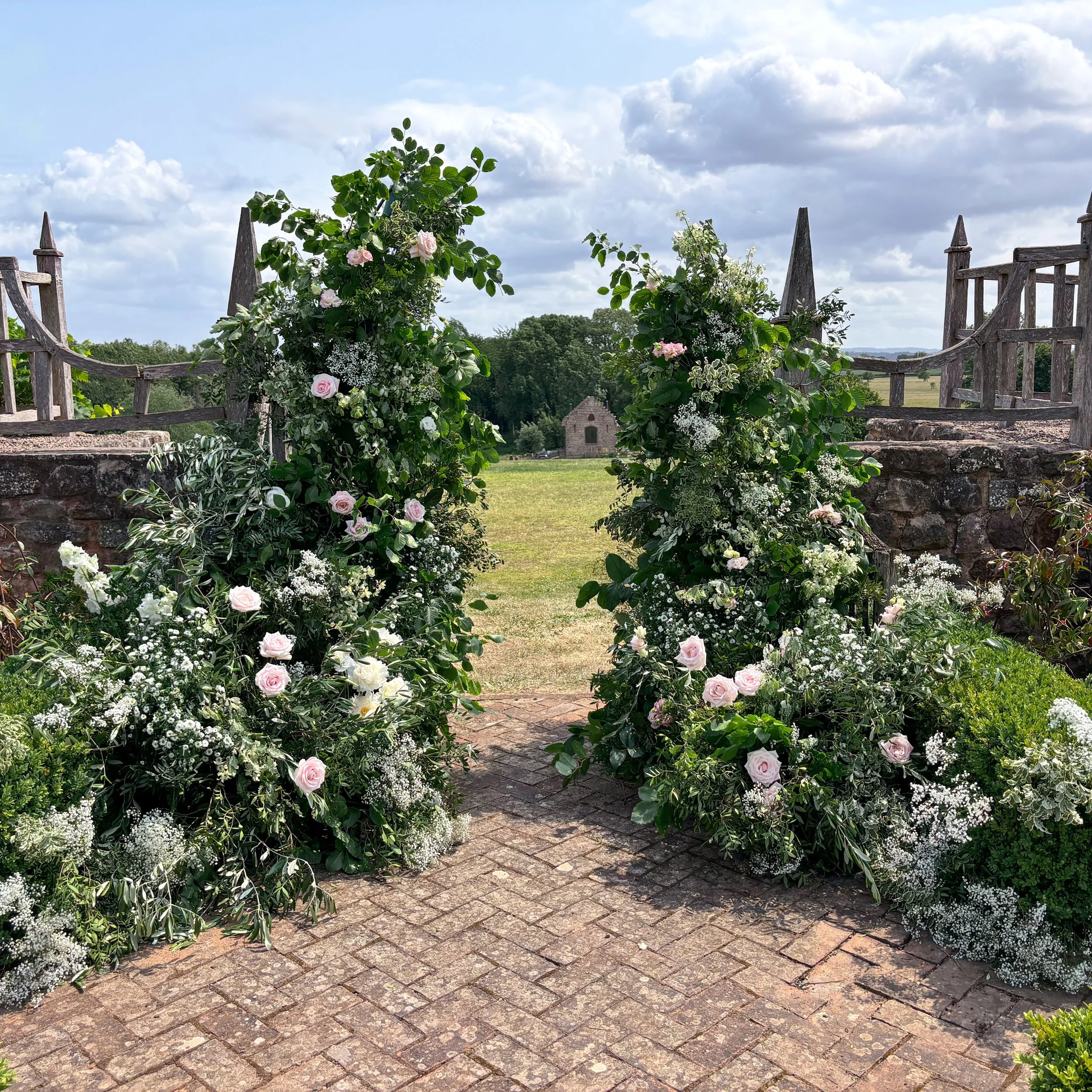 Wedding ceremony arch in the Sunken Garden at Pauntley Court, Gloucestershire, with soft pink and buttercream roses and abundant greenery.